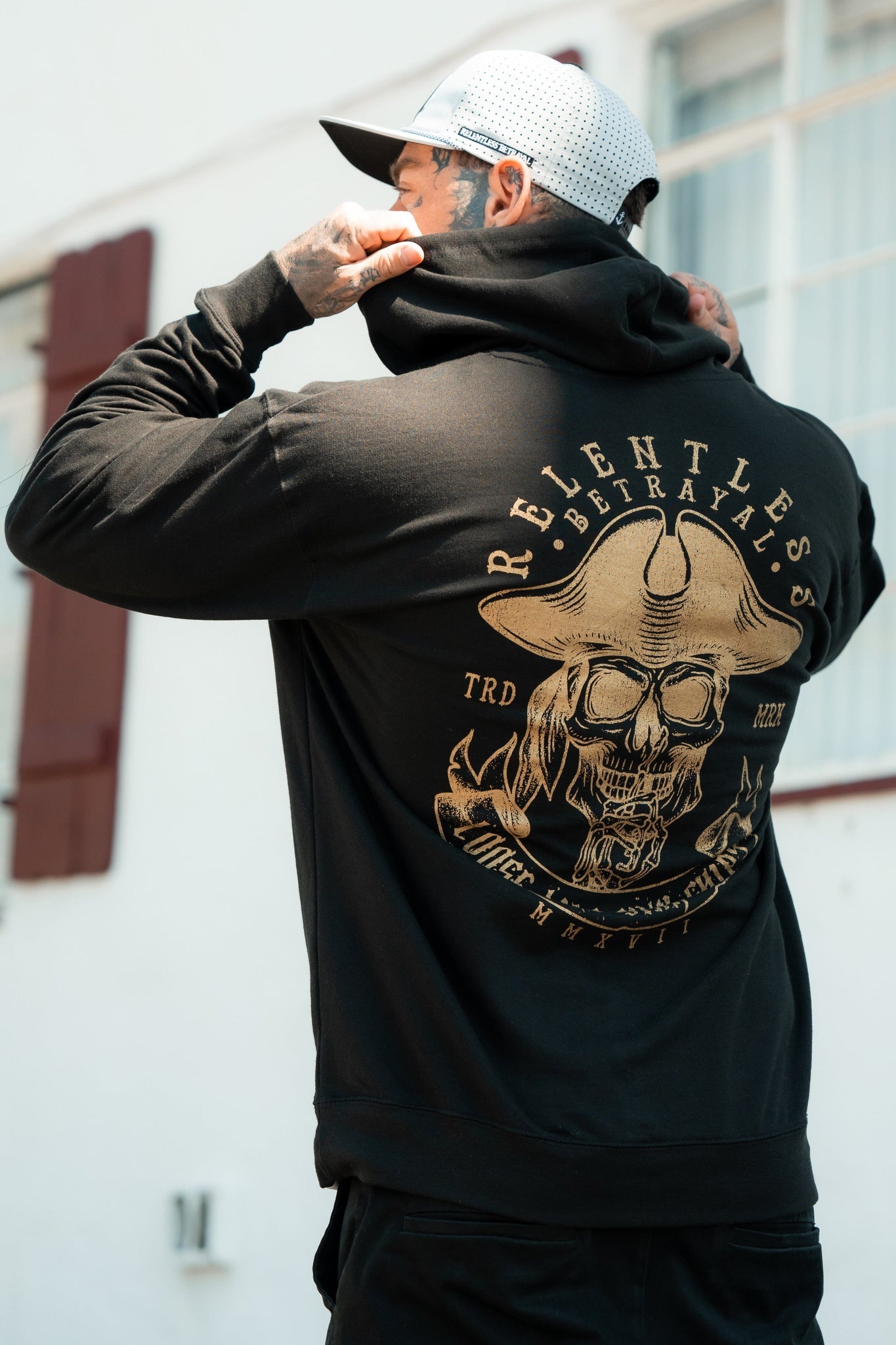 Outdoors near a building, someone adjusts the hood of their black Loose Lips Sink Ships Gold Foil Hoodie from Relentless Betrayal. The hoodie boasts an ornate skull and horns design with Relentless Betrayals on the back. They pair it with a white mesh cap. The hoodie is a comfy cotton/poly blend.