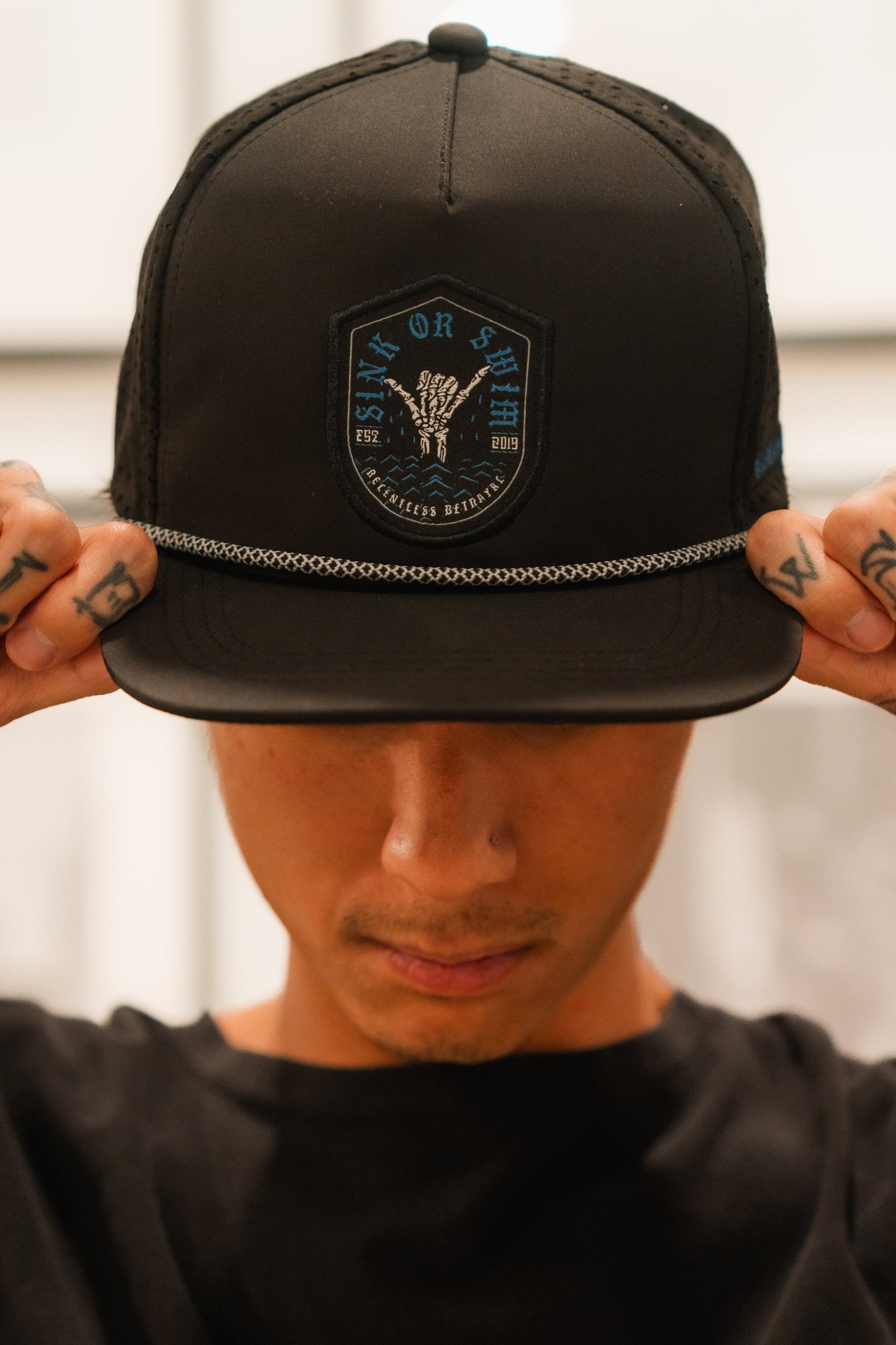A person adjusts their Sink Or Swim Snapback by Relentless Betrayal—featuring a detailed emblem and chain decoration—with both hands, uncovering tattoos symbolizing resilience. The blurred background highlights the caps design, embodying the spirit of perseverance amidst challenges.
