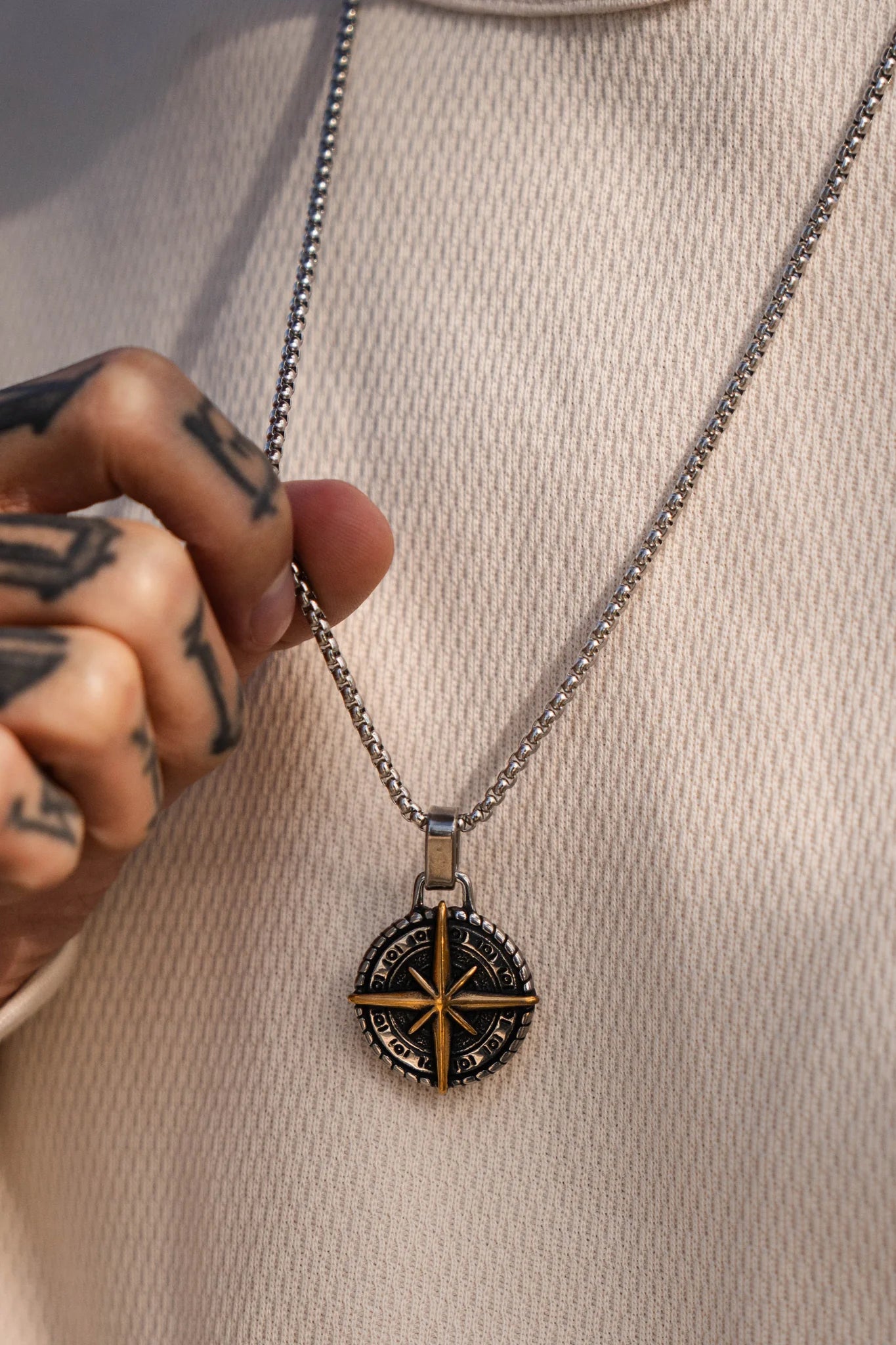 A tattooed person holds the Relentless Betrayal Polaris Golden Star Necklace, which has a circular compass pendant with a golden star at its center and sparkling gemstones. They wear a textured, light sweater that complements the necklaces elegance.