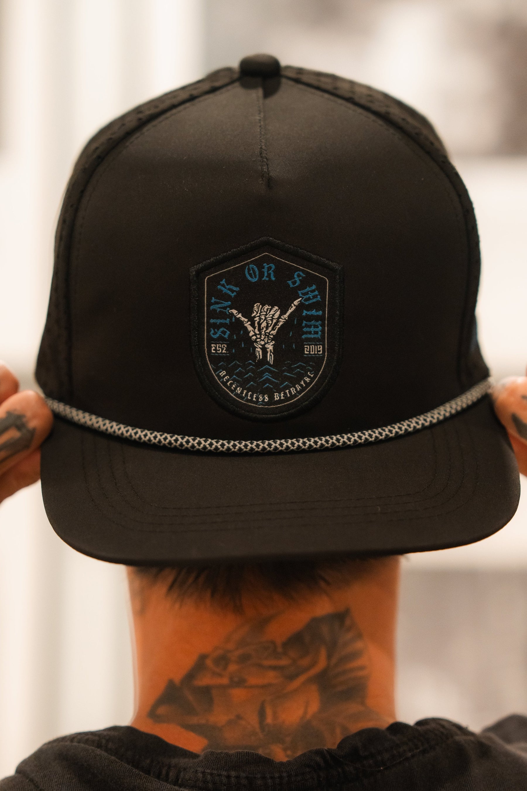 A tattooed individual adjusts their Relentless Betrayals Sink Or Swim Snapback cap, featuring a skeletal hand logo for resilience and a decorative rope above the brim, embodying the spirit of overcoming challenges.