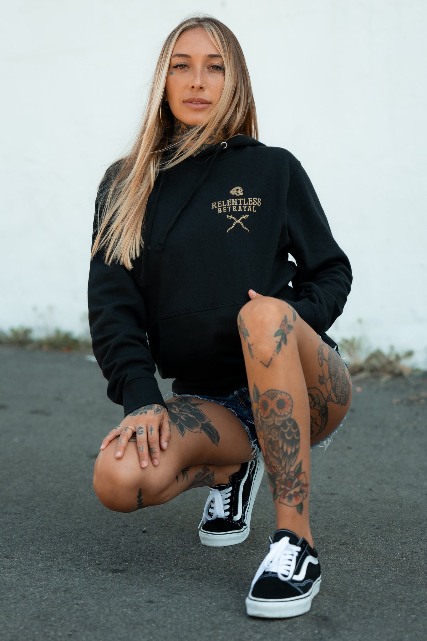 A person with long hair kneels on an outdoor pavement, wearing a Loose Lips Sink Ships Gold Foil Hoodie by Relentless Betrayal in a cotton/polyester blend. Its paired with denim shorts and black Vans, while prominent tattoos enhance their unique style.