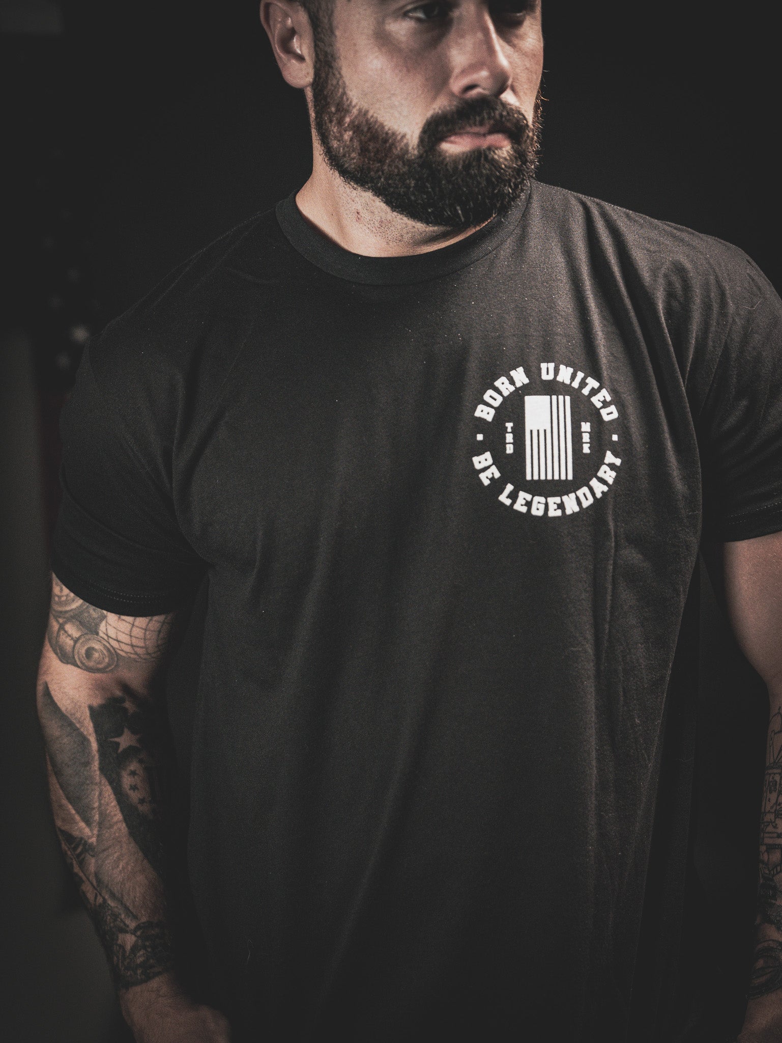 A man with a beard and tattoos on his arms is wearing a black T-shirt with the text Born United Be Legendary and a graphic design. The background is dark and the image is focused on the upper half of his body.