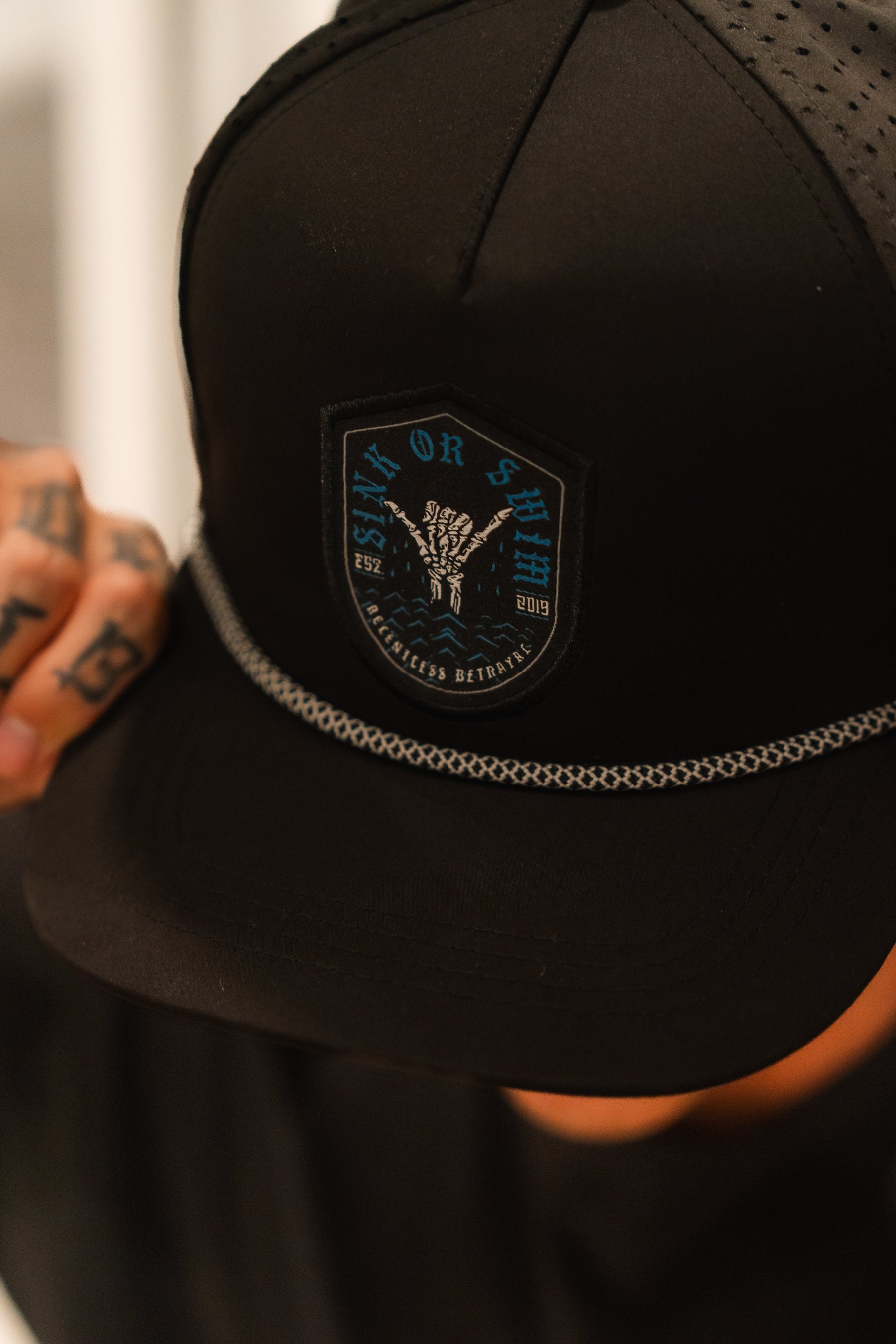 A person adjusts the brim of a black Sink Or Swim Snapback by Relentless Betrayal, adorned with a patch logo and chain detail, symbolizing resilience. Tattoos on their hand highlight their perseverance in overcoming challenges.