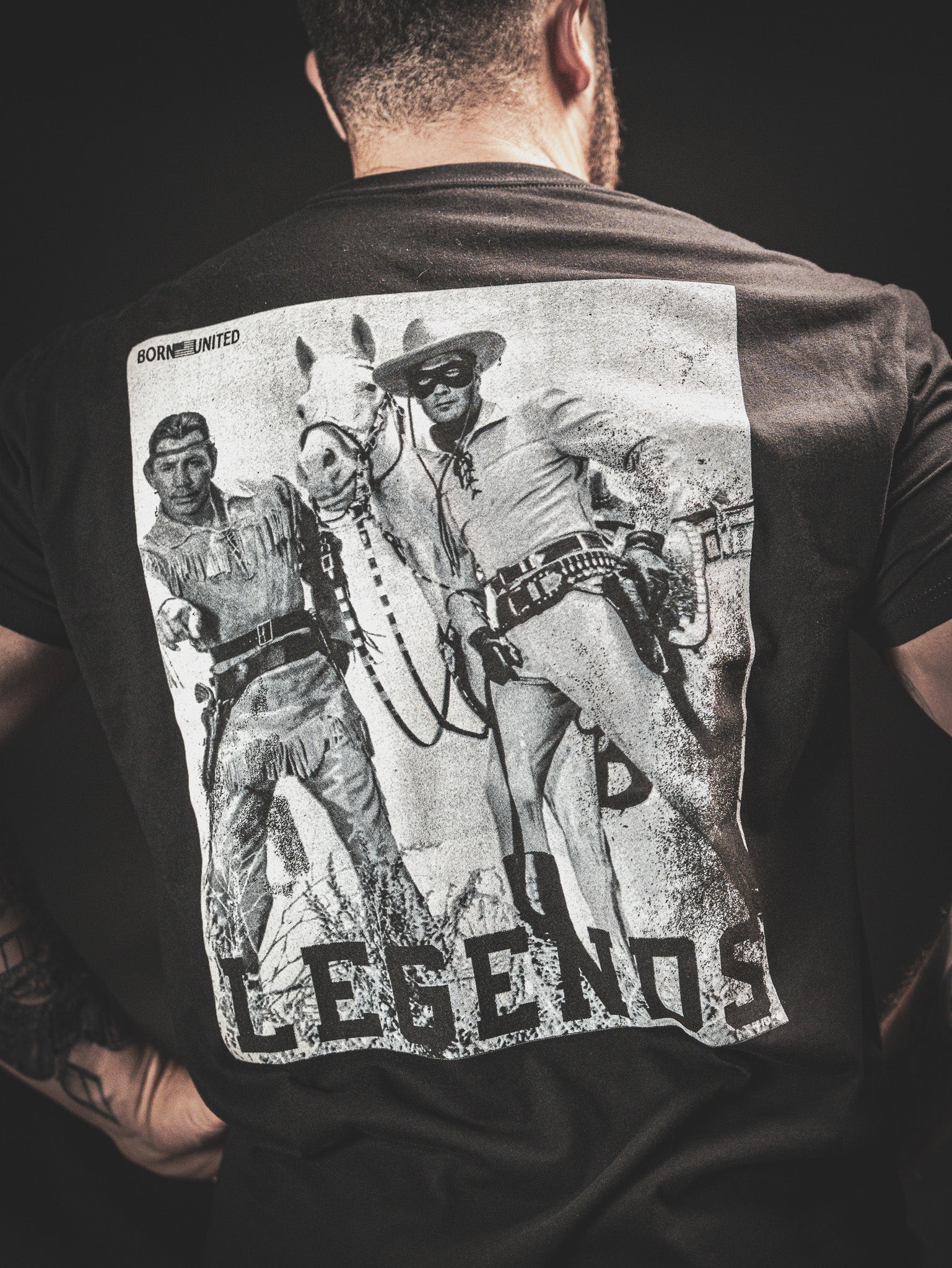 Back of a person wearing a black T-shirt featuring a vintage-style print of two cowboys from an old Western, alongside a horse. The word LEGENDS is printed at the bottom, and the word BORNLIMITED is above the image.