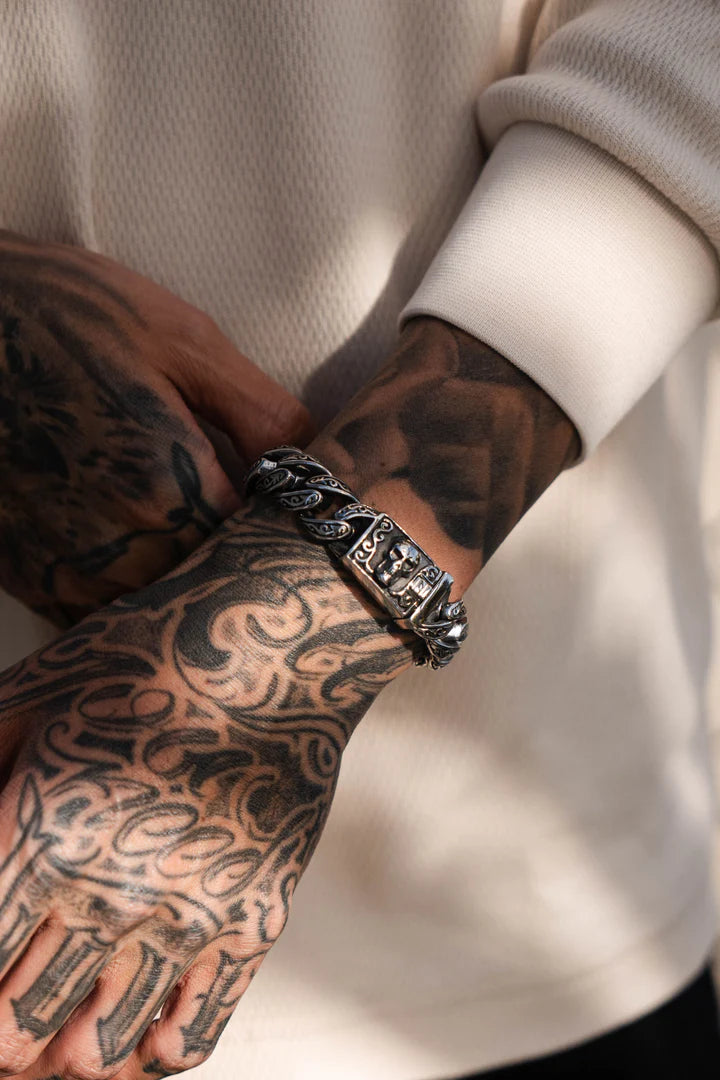 A person with tattooed hands wears a cream long-sleeve shirt and showcases the Deaths Door Bracelet by Relentless Betrayal, a captivating piece in stainless steel. The bracelets intricate designs are beautifully highlighted by the tattoos and its artistic placement.