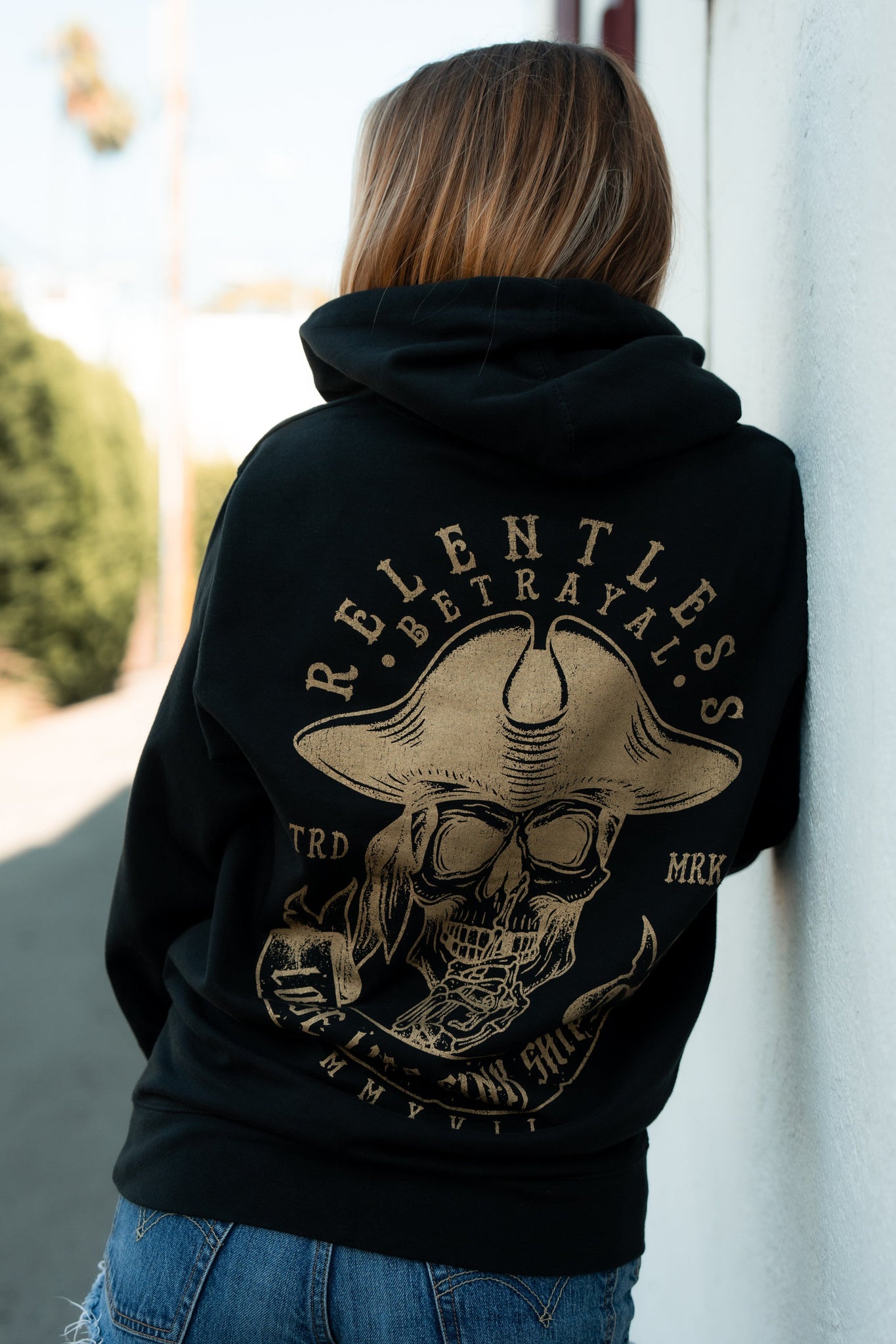 A person with long hair, wearing the Loose Lips Sink Ships Gold Foil Hoodie by Relentless Betrayal, stands against a white wall. This unisex black hoodie features a skull in a cowboy hat and is made from a comfy cotton/polyester blend, pairing effortlessly with their jeans.