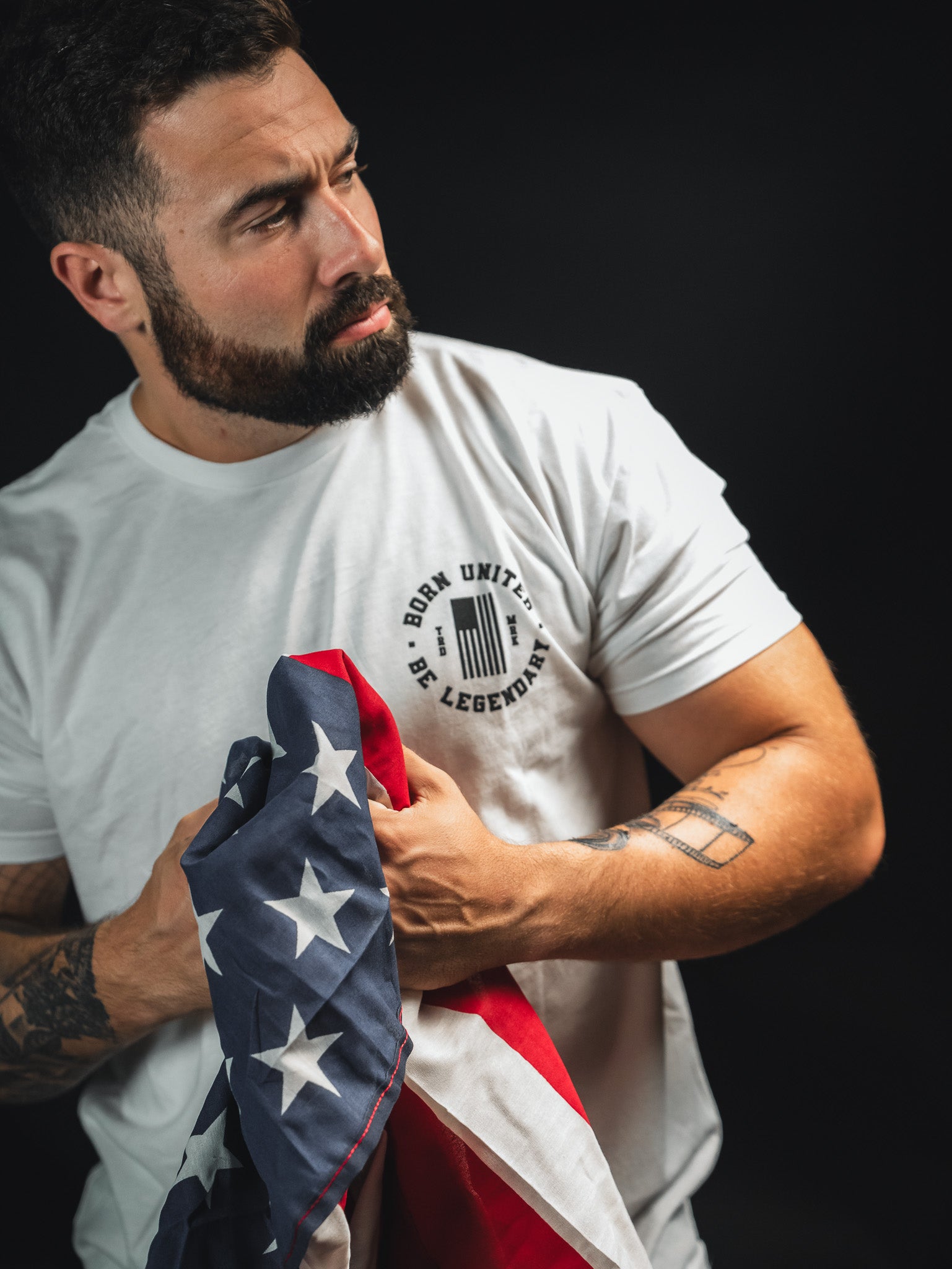 A man with a beard and tattoos holds a folded American flag. He is wearing a white T-shirt that reads BORN UNITED, BE LEGENDARY. The background is dark, highlighting the subject and the flag.