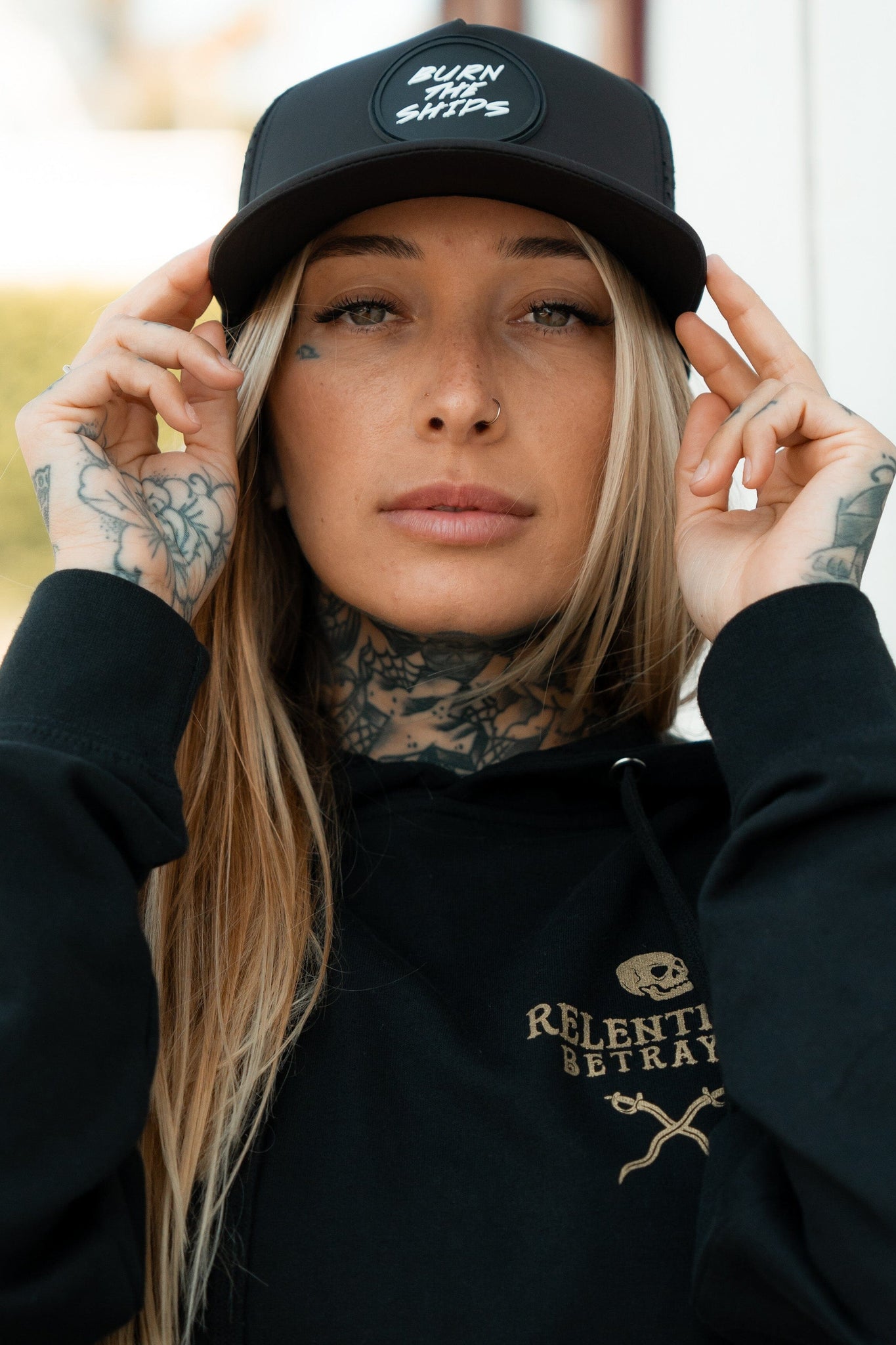 A person with long blonde hair, neck and hand tattoos, wears a Burn The Ships Black PVC Snapback by Relentless Betrayal. They have nose and lip piercings and are dressed in a black hoodie featuring RELENTLESS BETRAYAL, embodying the pirate lifestyle.