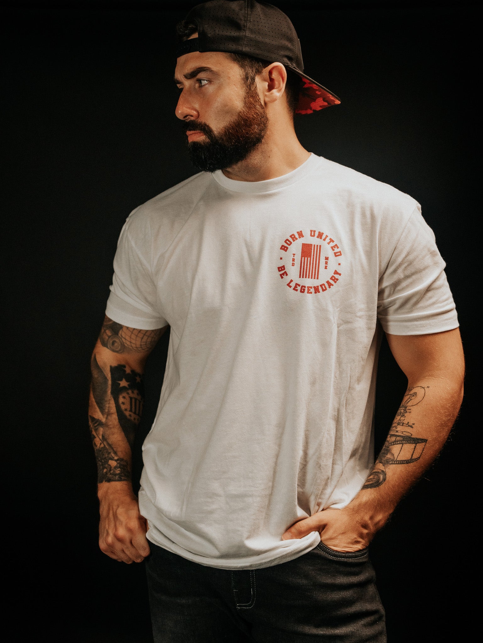A bearded man, wearing the Legends - Shrimp Co. white graphic t-shirt and a black cap from Born United Private Label, stands against a dim backdrop. His tattooed arms and serious leftward gaze create an enigmatic presence.