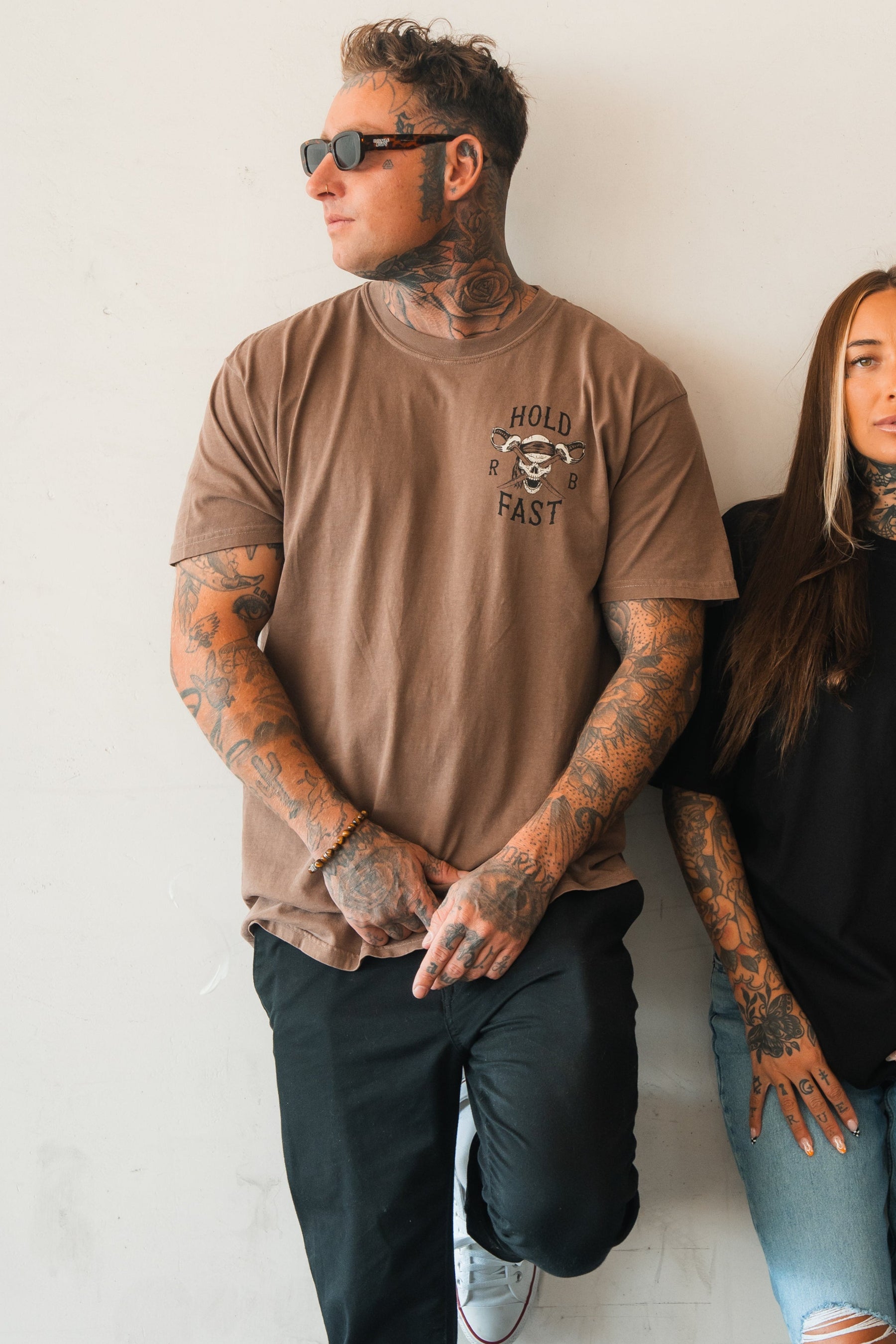 A tattooed man in sunglasses wears the Relentless Betrayal Hold Fast Premium Cocoa T-Shirt (heavyweight jersey cotton) with black pants, standing against a white wall with one leg up. A tattooed woman is partly visible beside him.