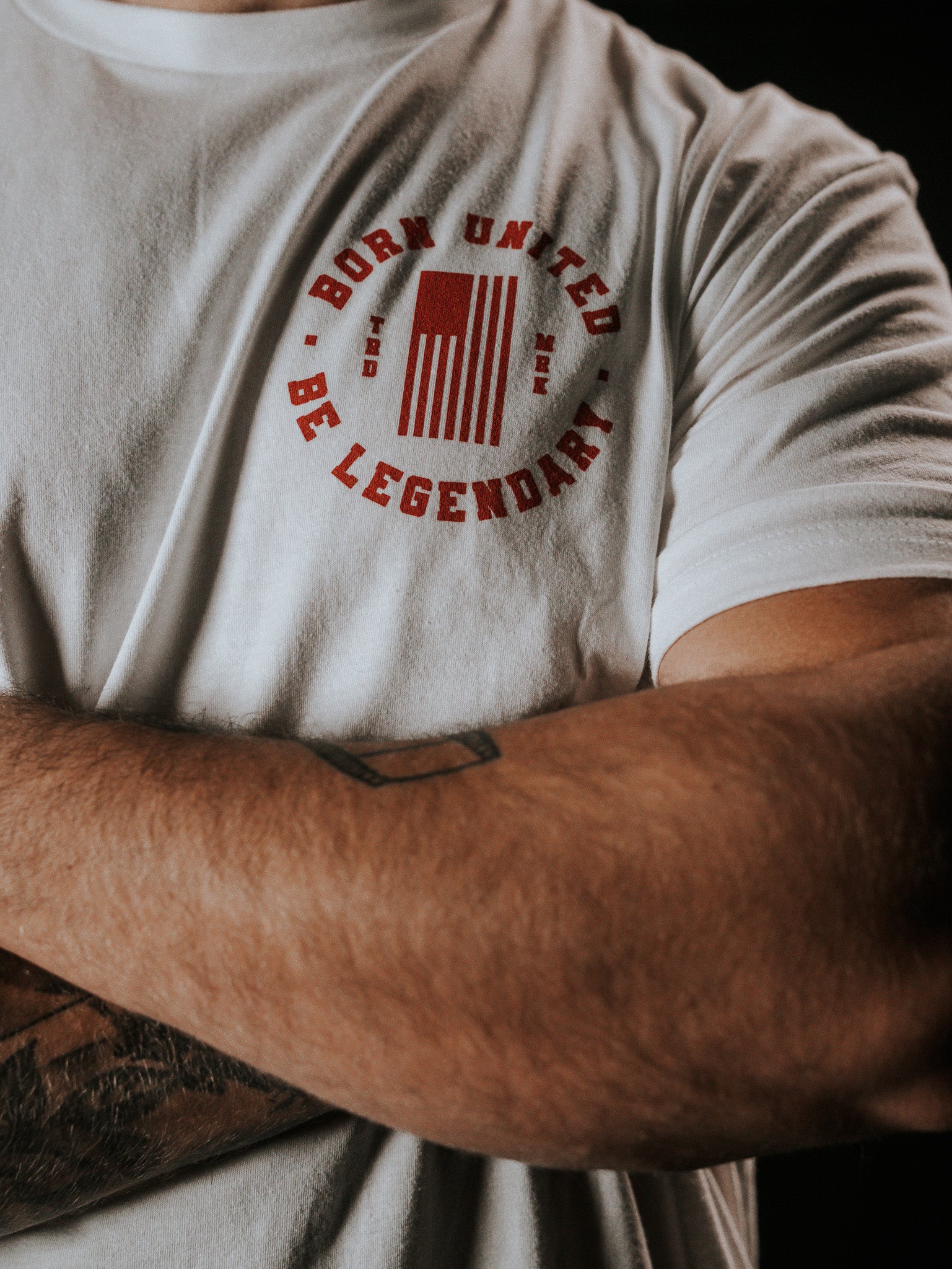 A person with crossed arms wearing a white T-shirt by Born United Private Label, featuring Born United and Be Legendary in red around a vertical American flag design, showcases vibrant arm tattoos.