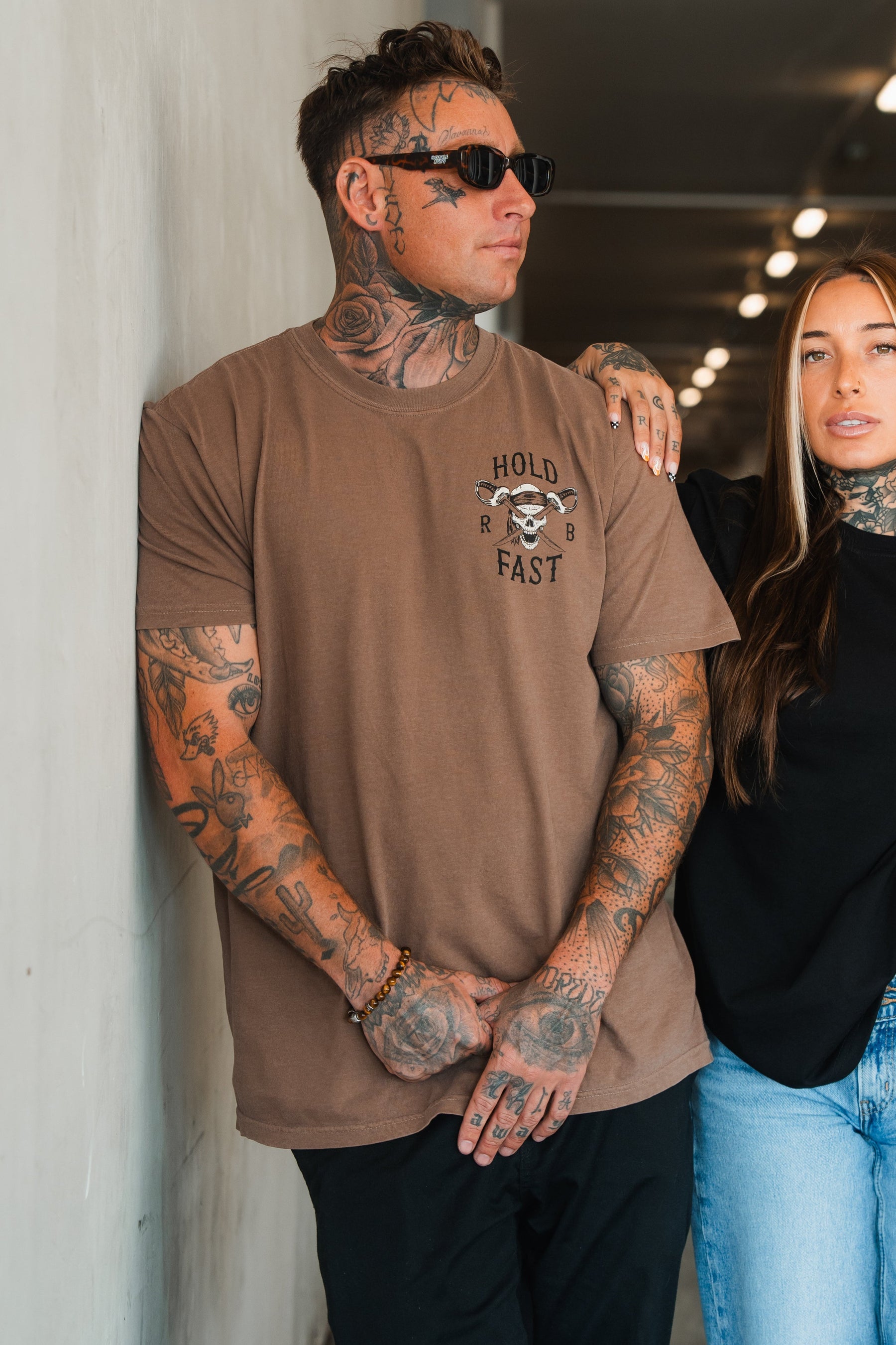 A tattooed man in sunglasses leans against a white wall, wearing the Relentless Betrayal Hold Fast Premium Cocoa T-Shirt. A tattooed woman with long brown hair stands beside him. Both look relaxed and casual.
