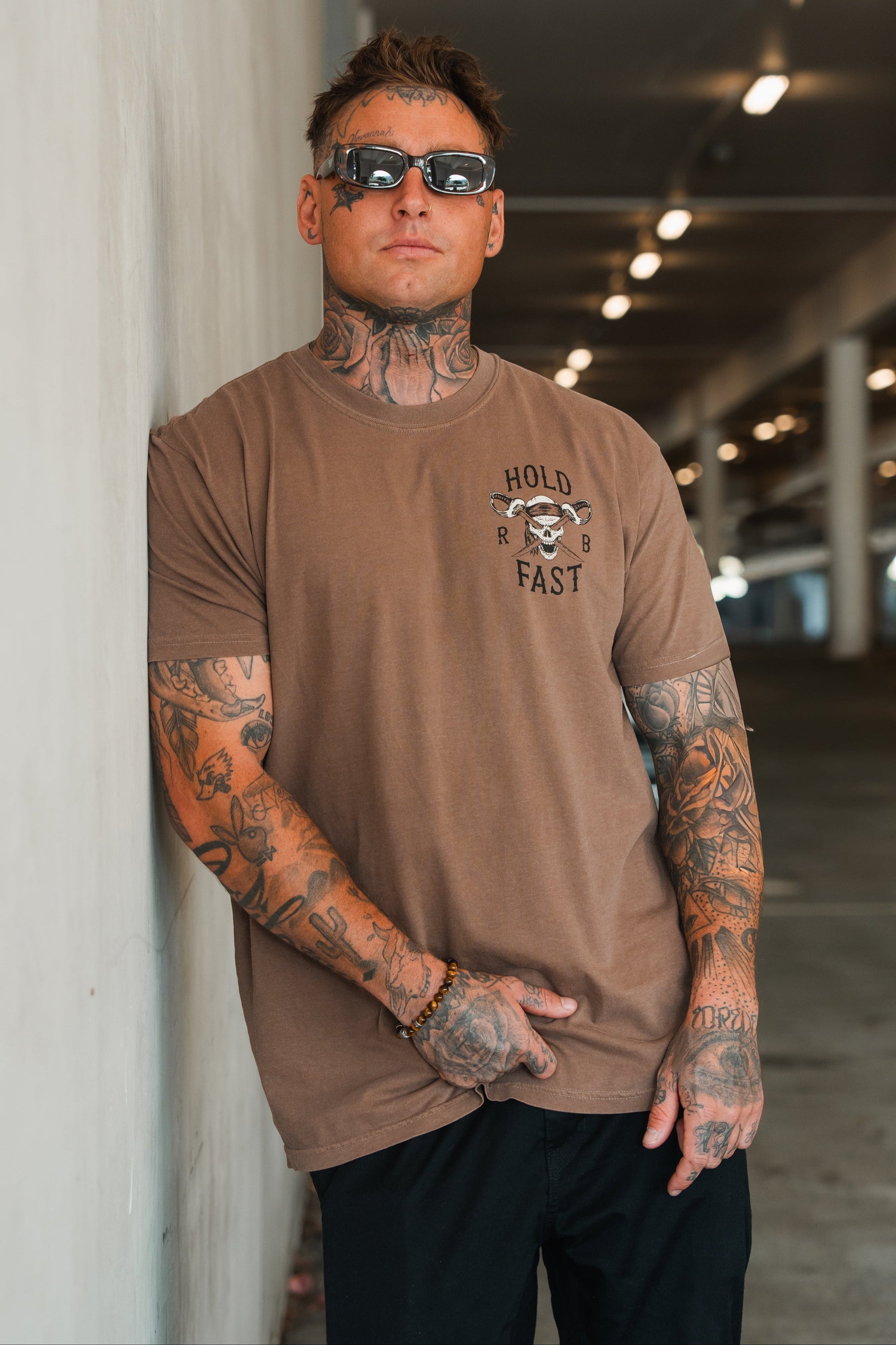 A tattooed man with short hair and sunglasses leans against a wall in a softly lit parking garage, wearing the Relentless Betrayal Hold Fast Premium Cocoa T-Shirt and black pants.