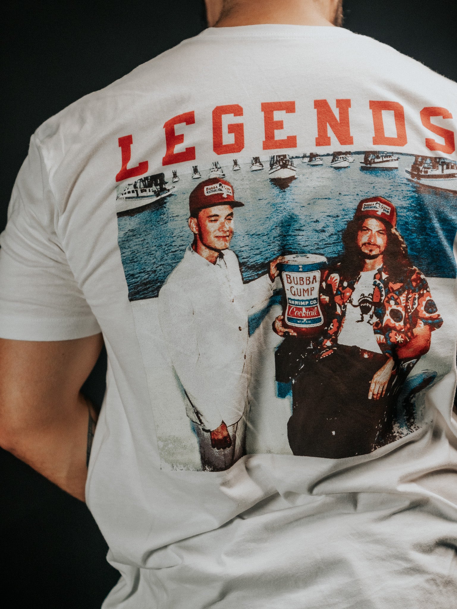 A person in a white Born United Private Label t-shirt featuring a vibrant back graphic from the Legends - Shrimp Co. collection: two iconic figures holding a Bubba Gump Shrimp Co. can above LEGENDS in bold red, against a lively waterfront with boats swaying gently.