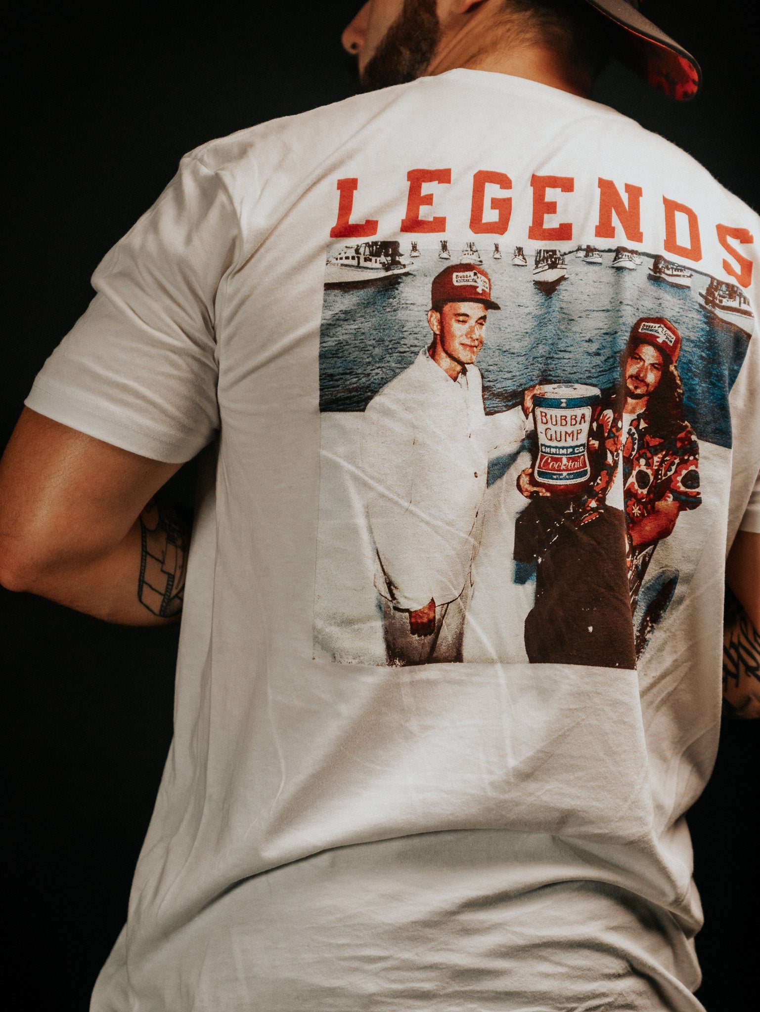 A man wears a Born United Private Label baseball cap and a white Legends - Shrimp Co. T-shirt, featuring a vibrant back image with three people by boats, one holding a Bubba Gump box, and LEGENDS prominently displayed in bold red letters.