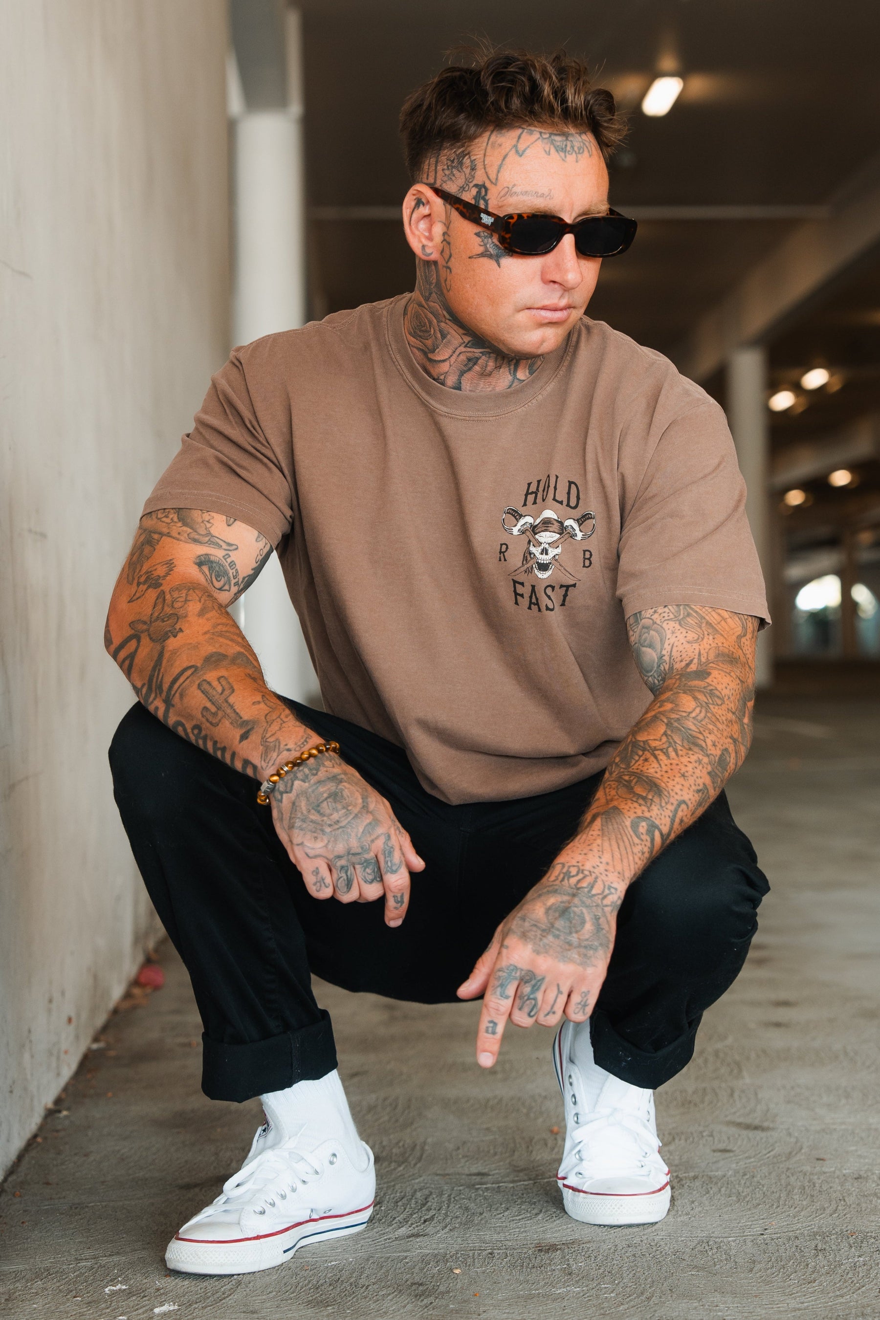 A tattooed man in a Relentless Betrayal Hold Fast Premium Cocoa T-Shirt, black pants, white sneakers, and sunglasses crouches in a parking garage, looking to the side with one hand on his knee and the other pointing downward.