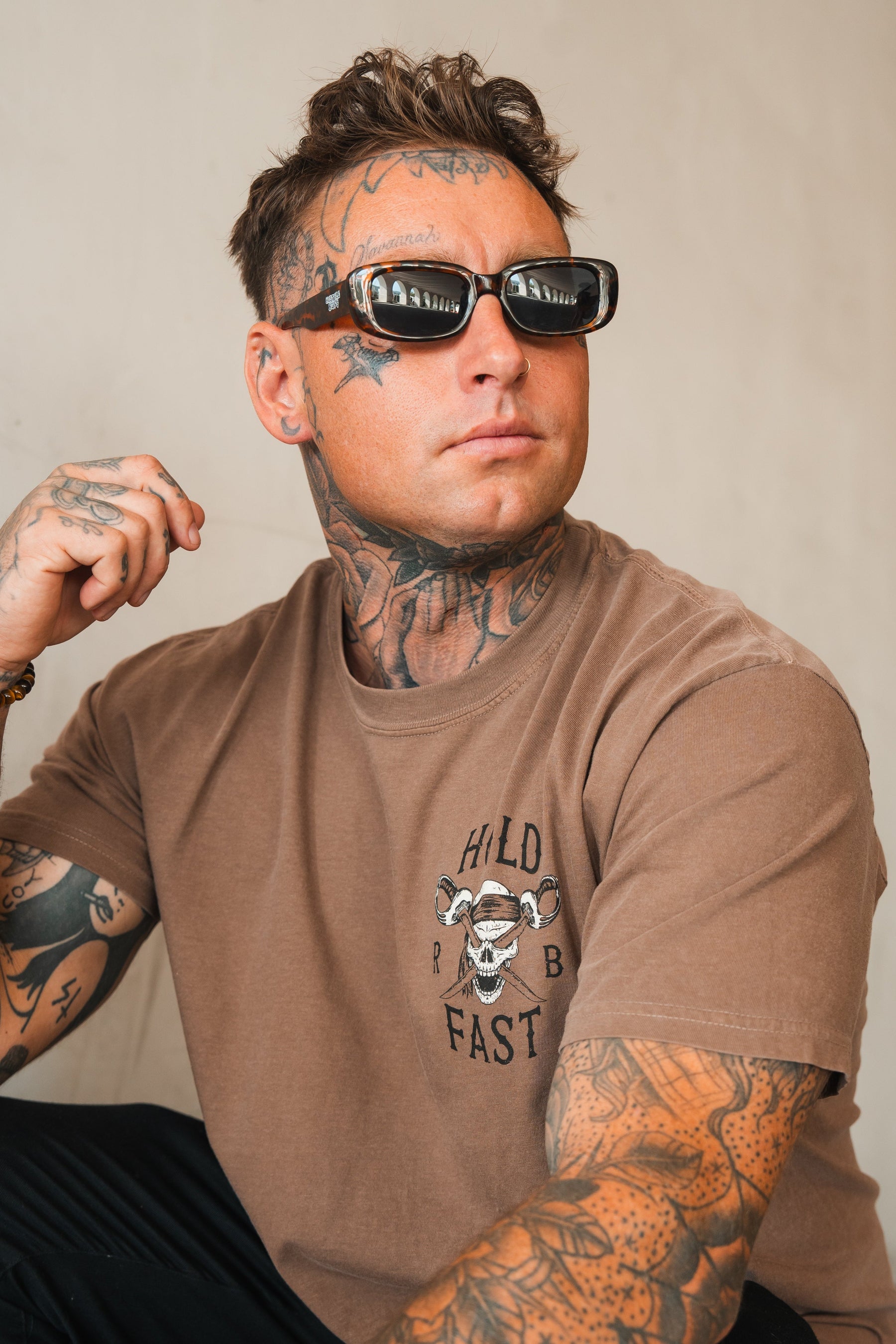 A tattooed man in sunglasses wears a Relentless Betrayal Hold Fast Premium Cocoa T-Shirt with a skull graphic. He looks to the side with a neutral expression against a plain background.