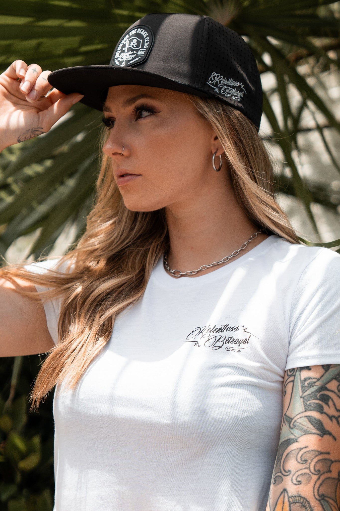 A tattooed woman wears a white T-shirt and adjusts her Relentless Betrayal Black Flag Snapback cap while standing outside against a lush, green backdrop.