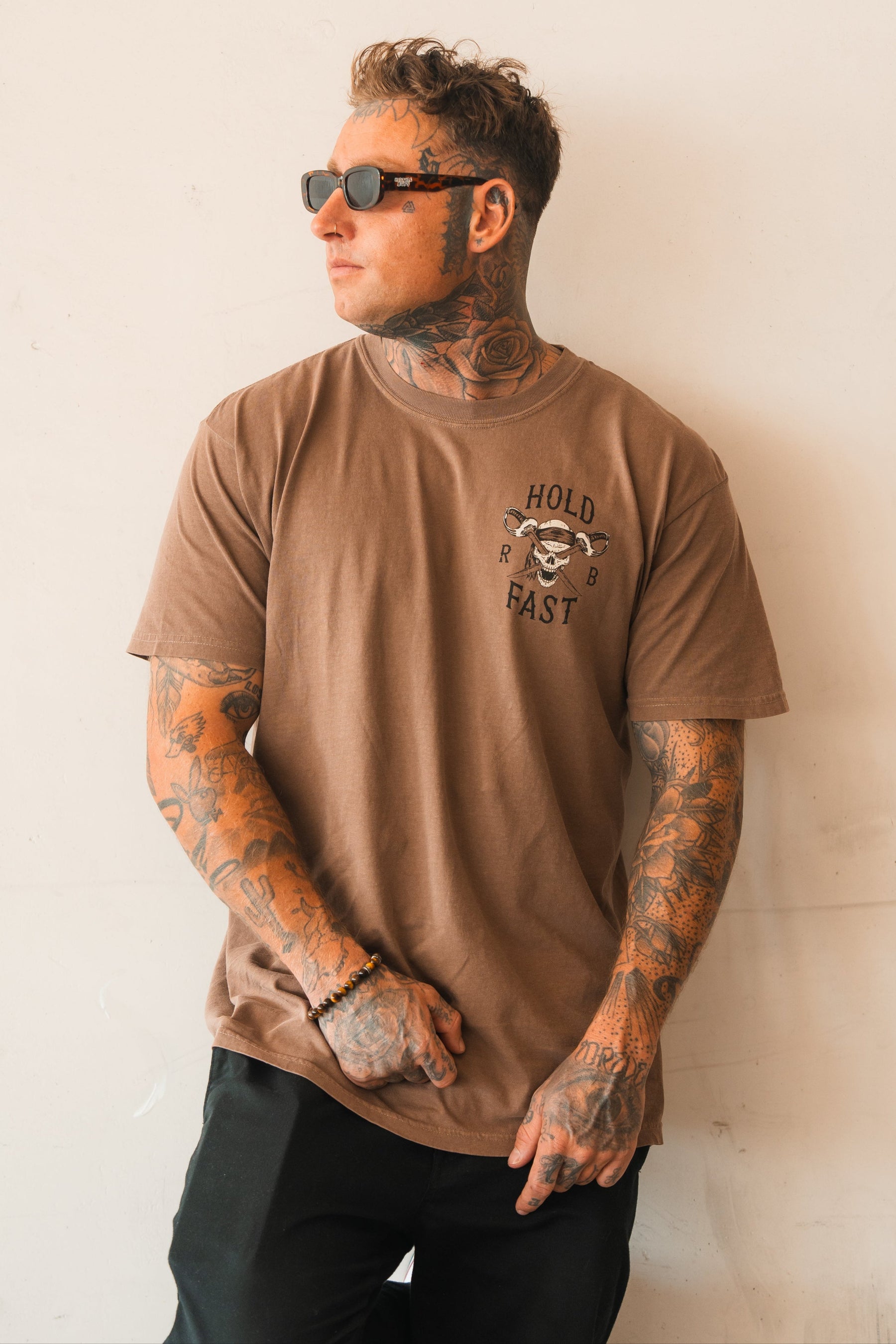 A tattooed man in sunglasses wears a Relentless Betrayal Hold Fast Premium Cocoa T-Shirt with a skull graphic, standing against a light wall and looking to the side while holding his shirt.