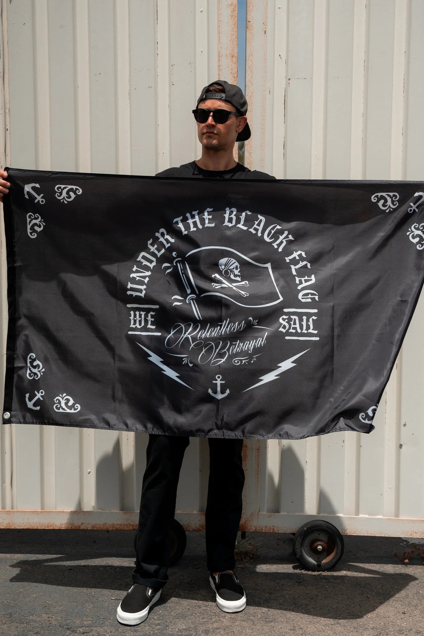 A person in a backwards hat and sunglasses holds Relentless Betrayals Under The Black Flag We Sail polyester flag with durable metal grommets against a light corrugated metal wall, featuring text and decorative symbols.