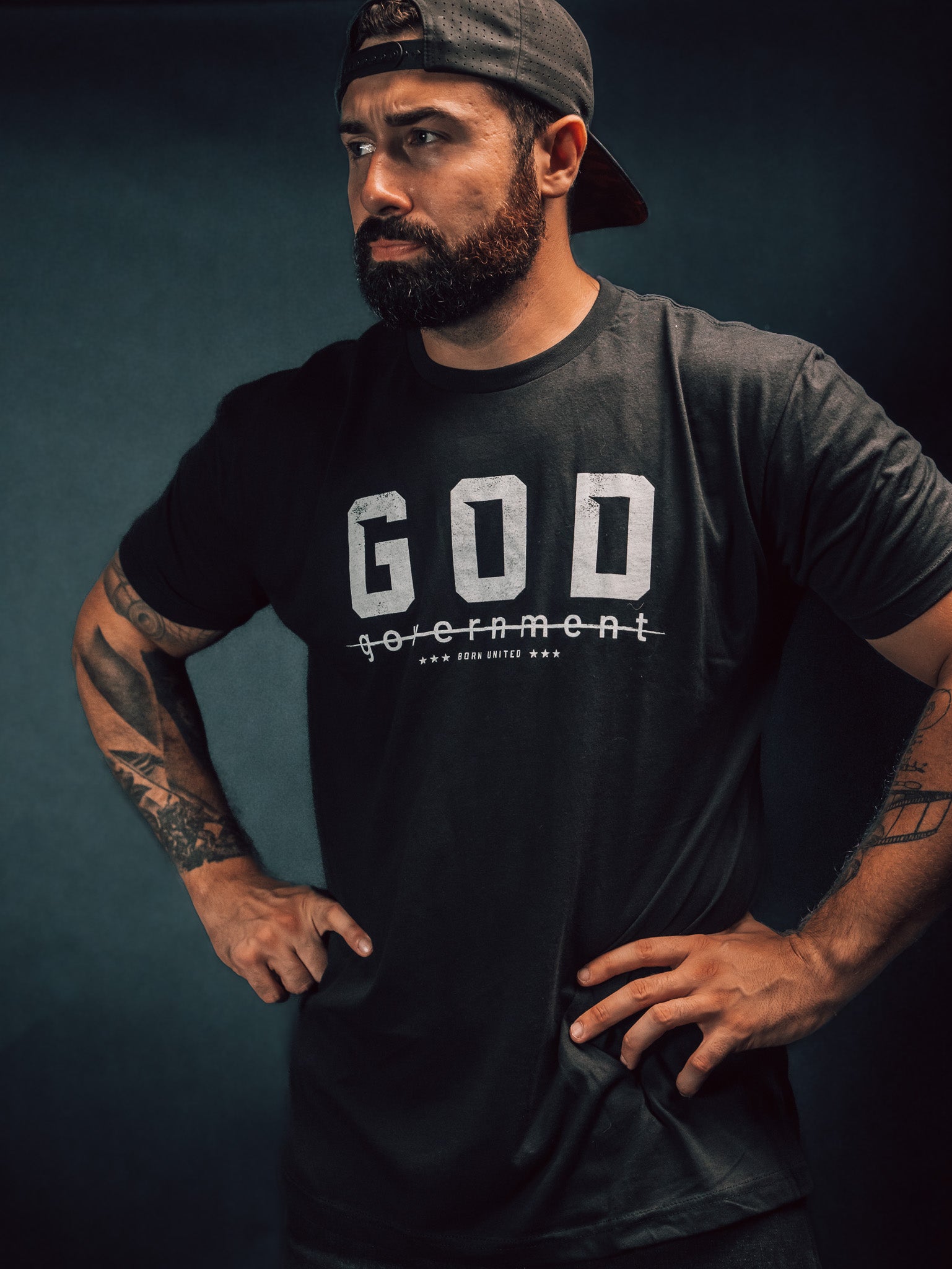 A bearded man, gazing to the side against a dark background, stands confidently with hands on hips. Hes wearing a reversed baseball cap and Born United Private Labels God Over Government t-shirt, which features the word government crossed out below GOD, challenging authority with his defiant posture.
