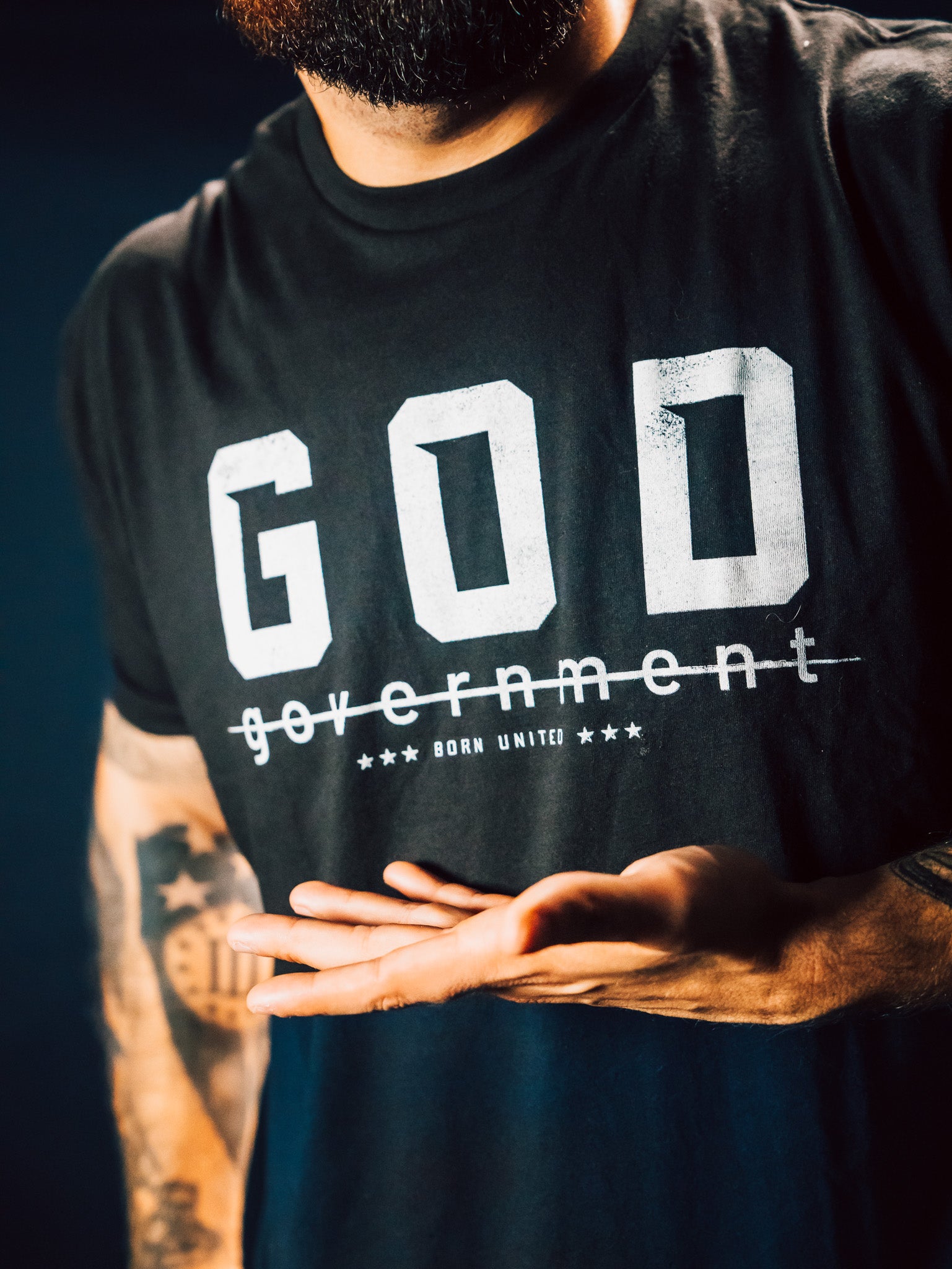 A person wearing a God Over Government black T-shirt by Born United Private Label, featuring stars and bold text, stands confidently in a dark background. Their tattooed arm gestures respect for rulers and good conduct.