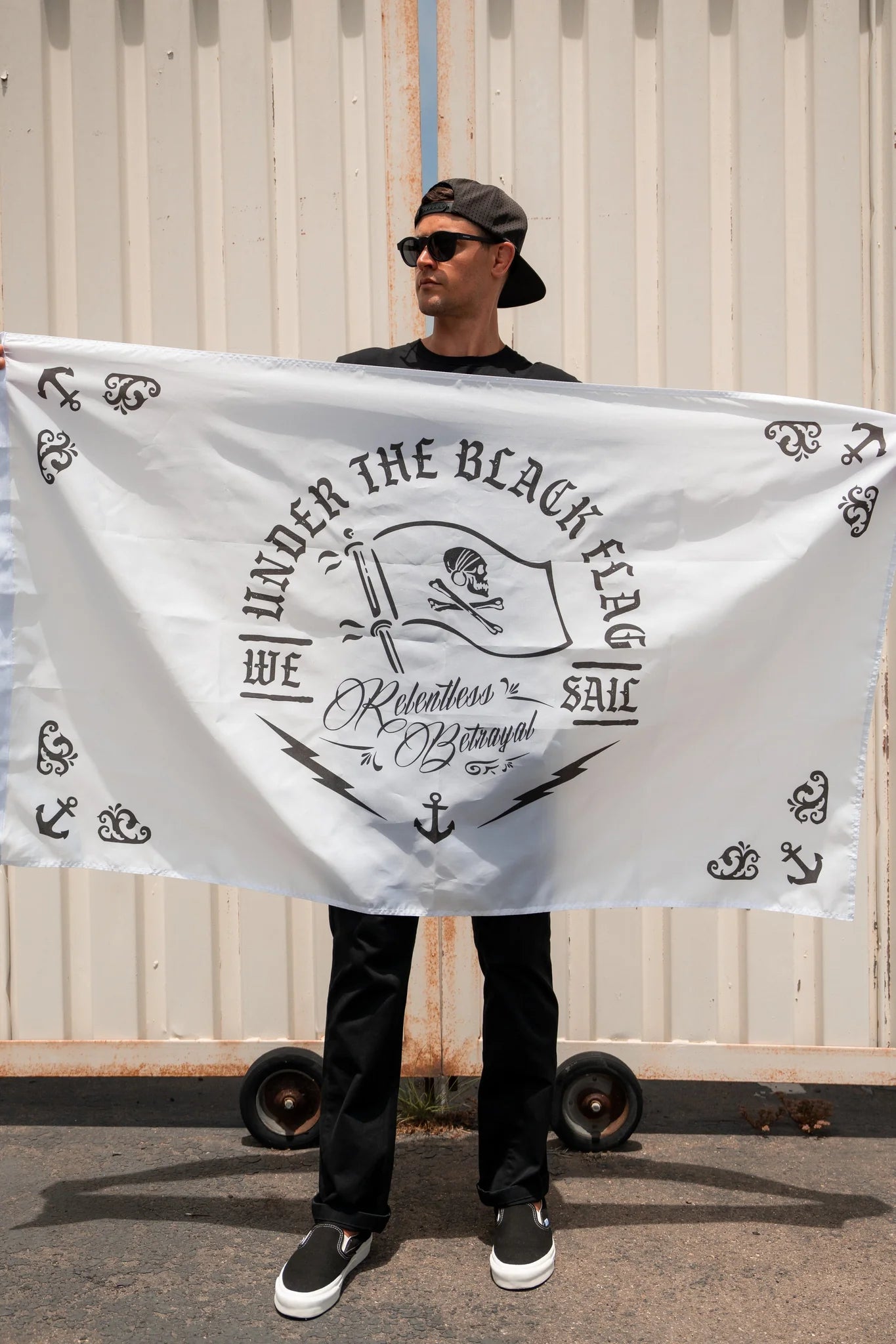 A man confidently stands against a corrugated metal background, holding the Under The Black Flag We Sail V2 Flag by Relentless Betrayal, crafted from durable polyester with nautical symbols and text. He wears a black cap, sunglasses, and black clothing.