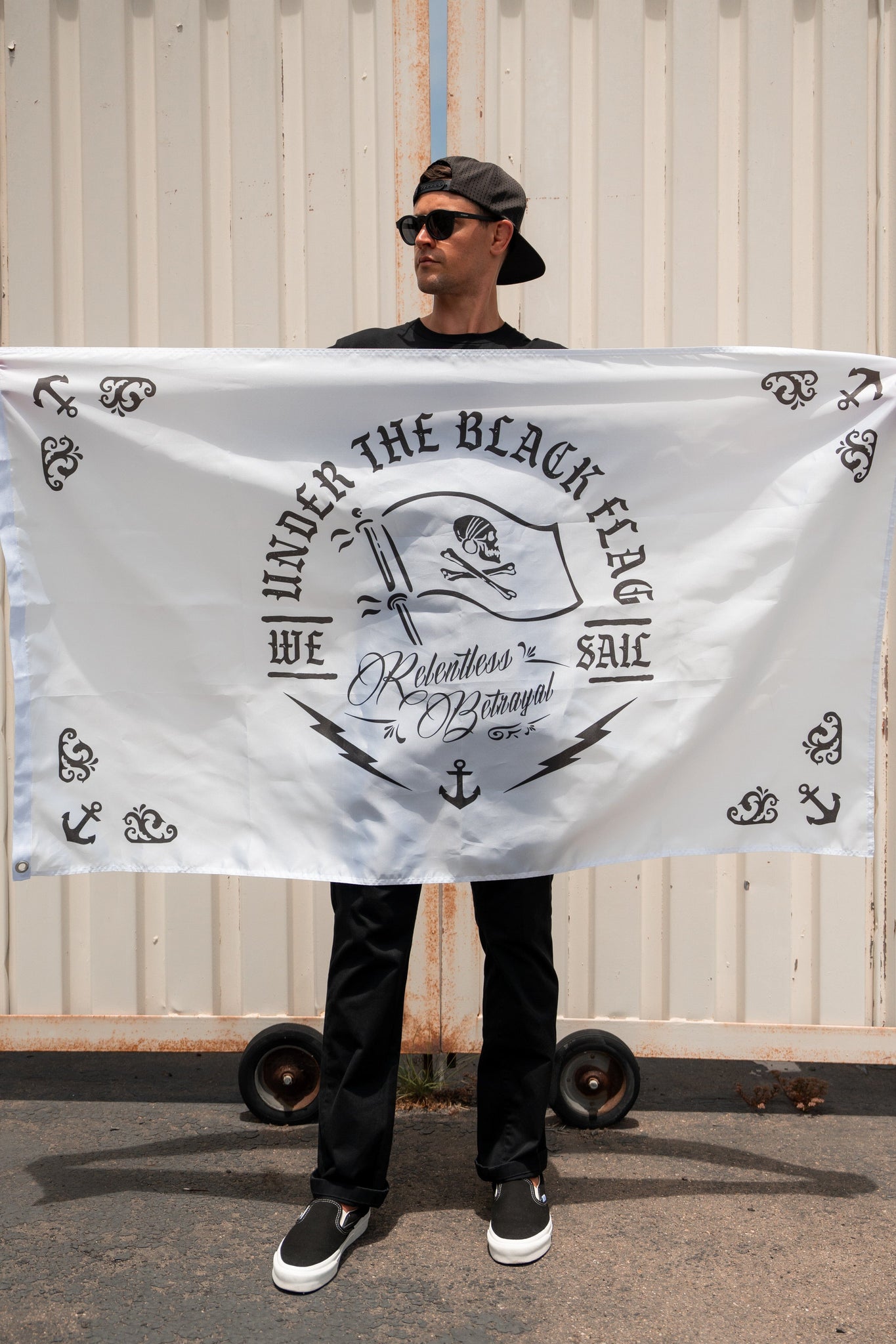 A man in a black outfit holds the durable polyester Under The Black Flag We Sail V2 Flag by Relentless Betrayal, featuring nautical designs and metal grommets, against a corrugated metal backdrop.