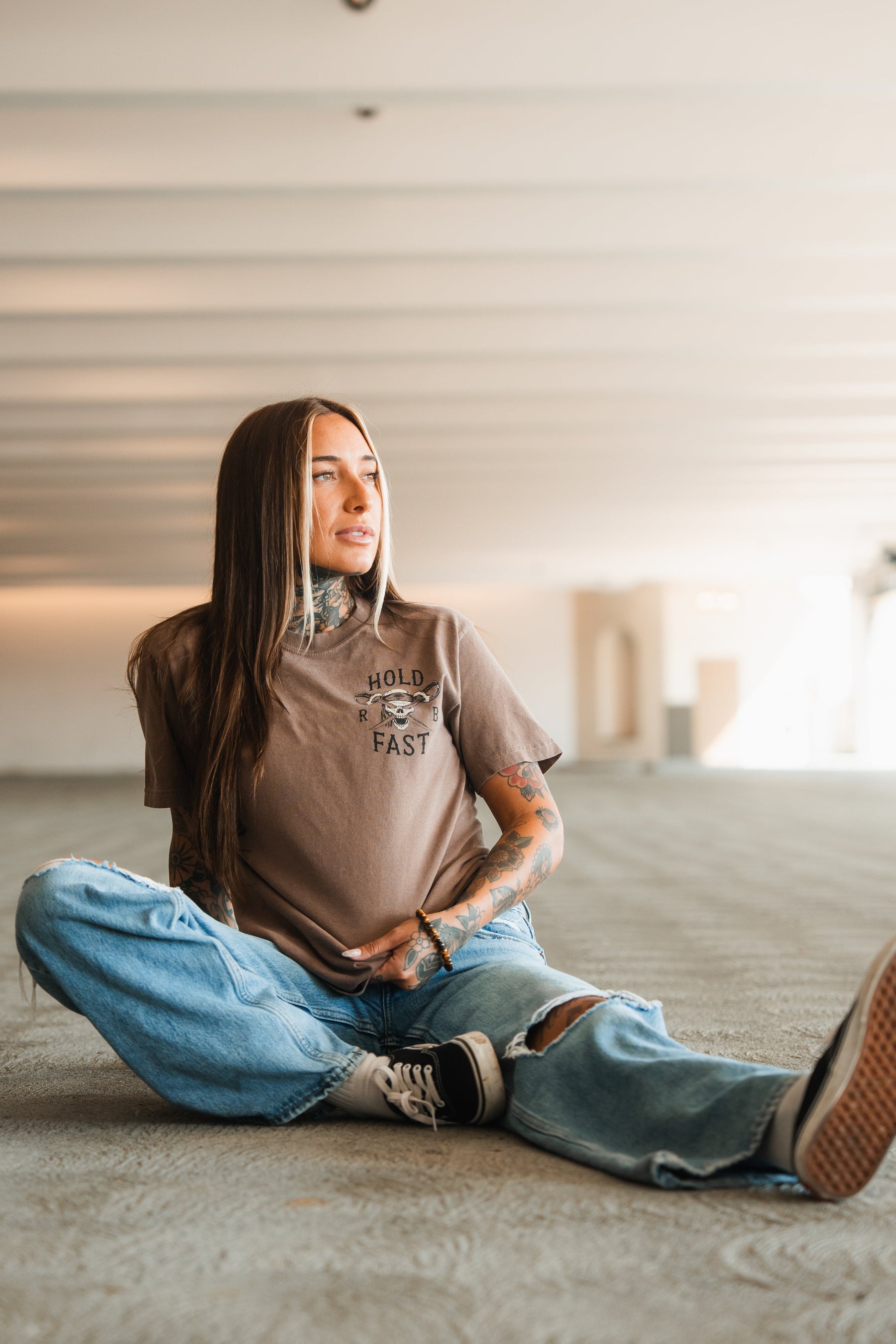 A person with long hair and tattoos sits casually on the floor of a spacious, well-lit room, wearing the Relentless Betrayal Hold Fast Premium Cocoa T-Shirt, ripped blue jeans, and sneakers, looking off to the side and smiling slightly.
