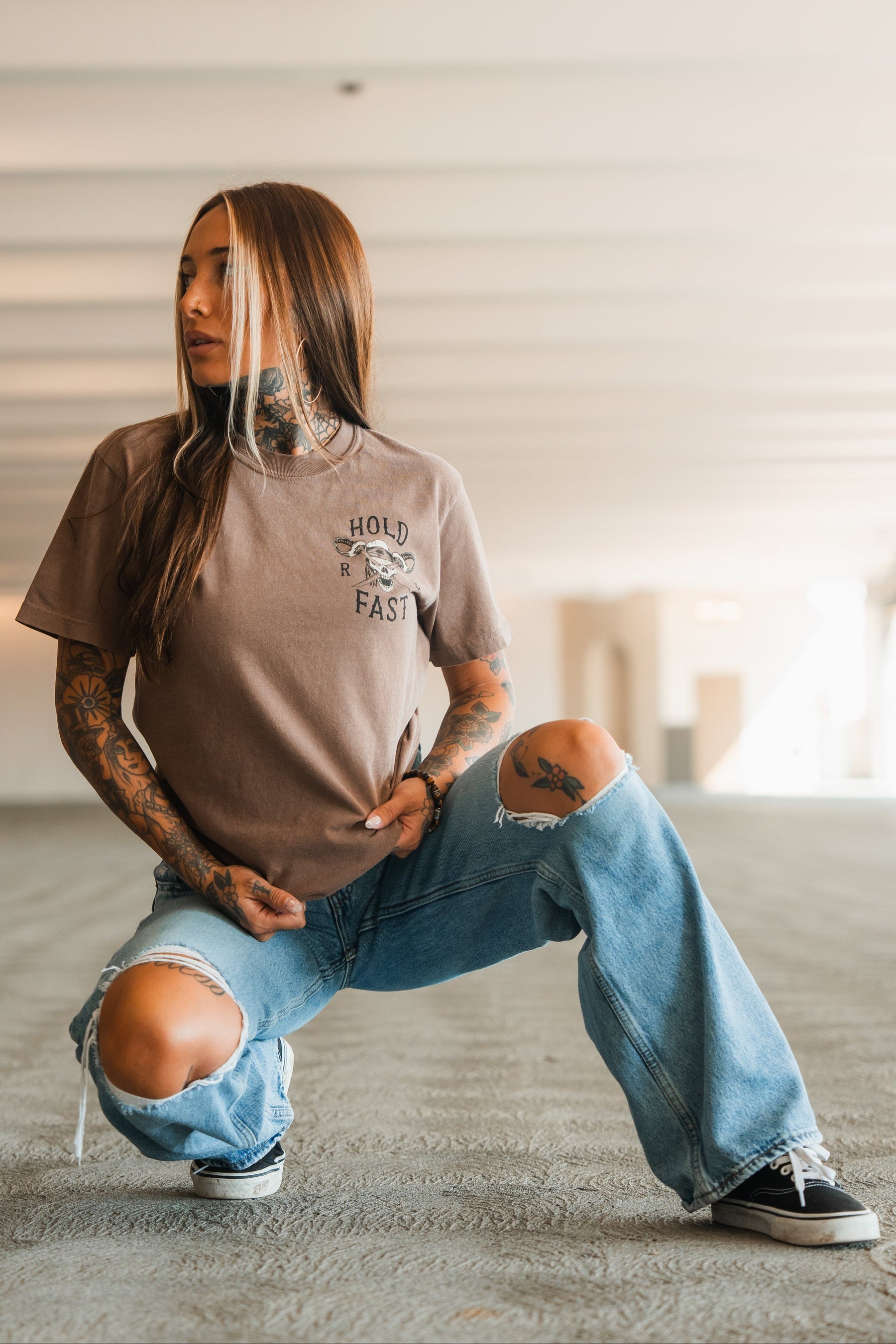 A woman with tattoos and long brown hair kneels on one knee in a bright, empty space, wearing a Relentless Betrayal Hold Fast Premium Cocoa T-Shirt, ripped blue jeans, and black sneakers, looking to the side with one hand on her shirt.