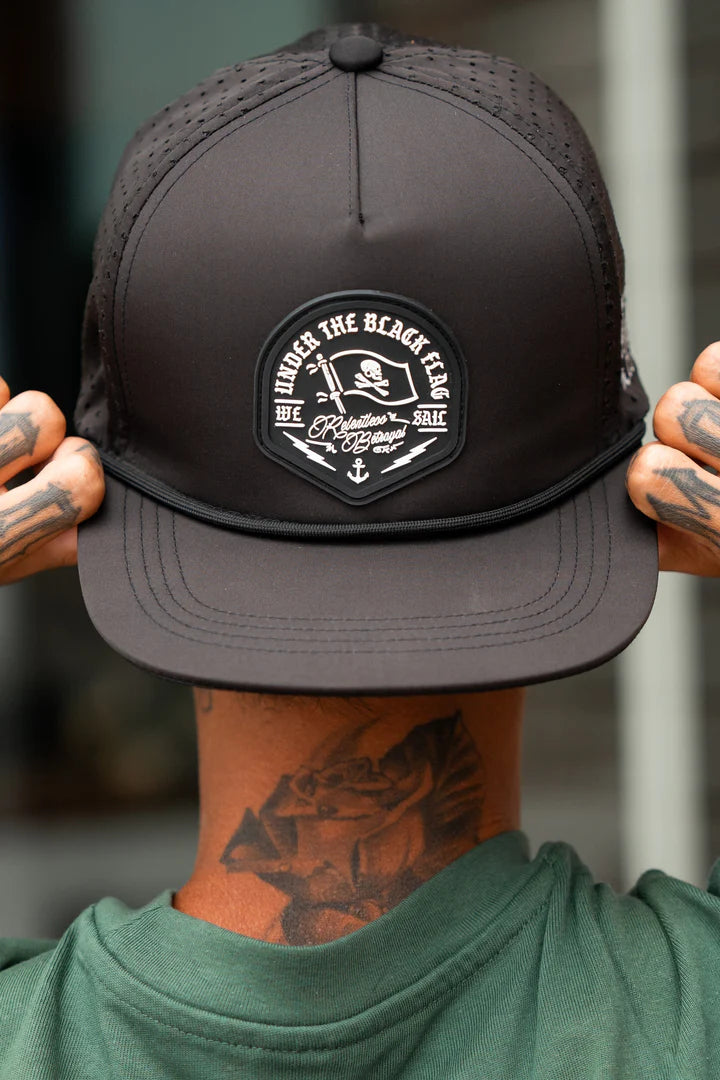 Facing away, the person wears a Black Flag Snapback- Black cap from Relentless Betrayal with a pirate skull and crossbones patch reading Under the Black Flag We Sail. They embrace the pirate life with hand and neck tattoos, paired with a green shirt.