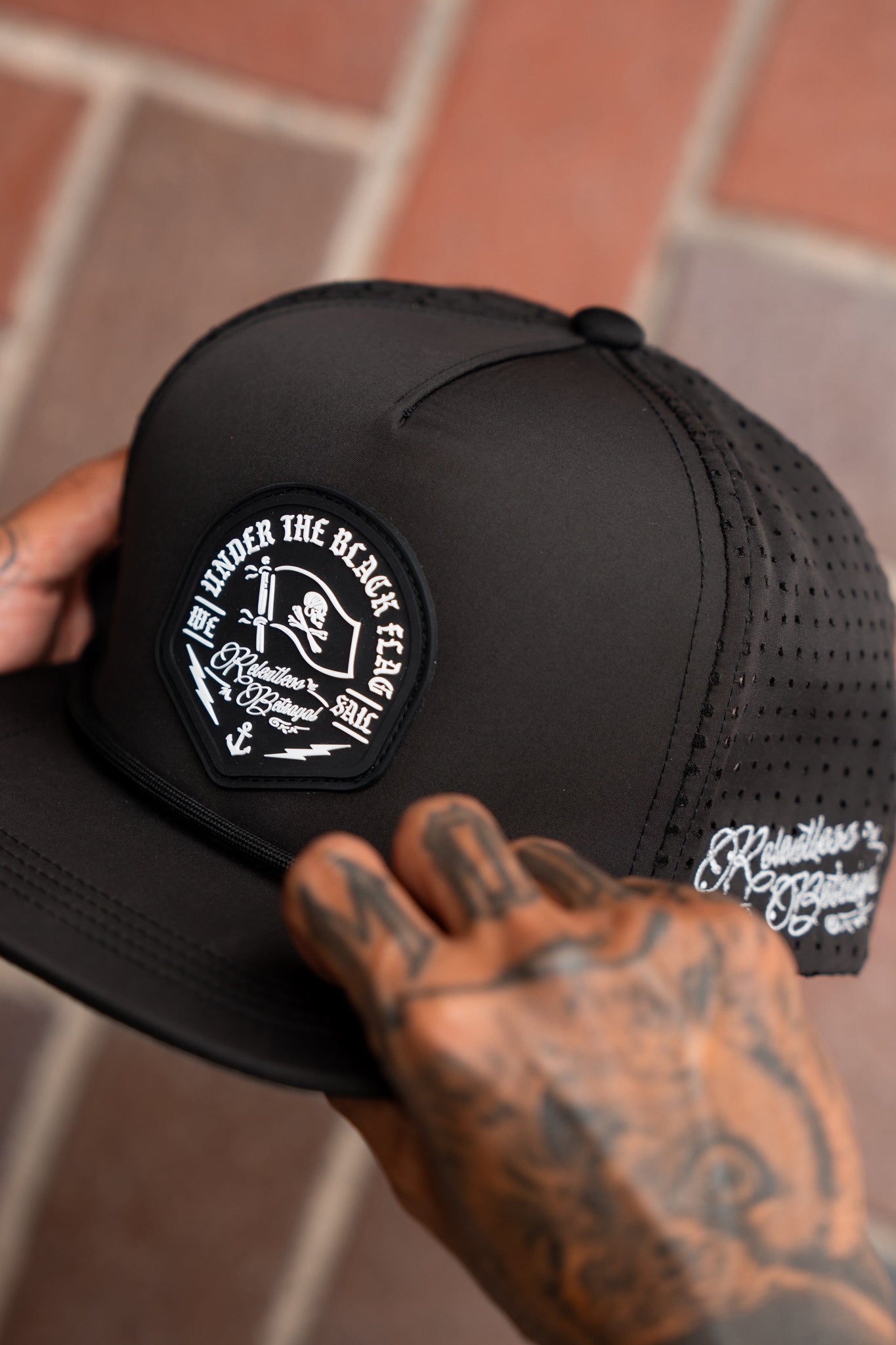 A person with tattooed hands adjusts a Relentless Betrayals Black Flag Snapback- Black hat, which features a nautical-themed patch with an anchor, sword, skull, and Under the Black Sea. This water-resistant trucker hat has a snapback closure and white embroidered text on the side.