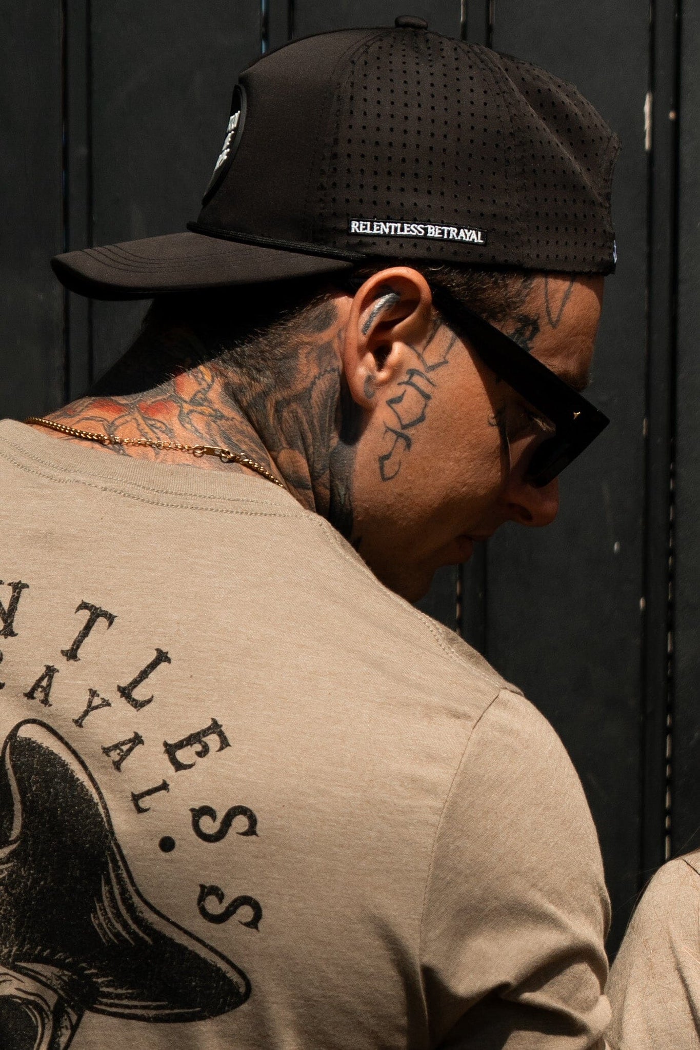 A person in a black snapback, sunglasses, and a beige shirt with a back design gazes sideways, their neck adorned with tattoos. The cap features Relentless Betrayal, echoing pirate-like mystery.