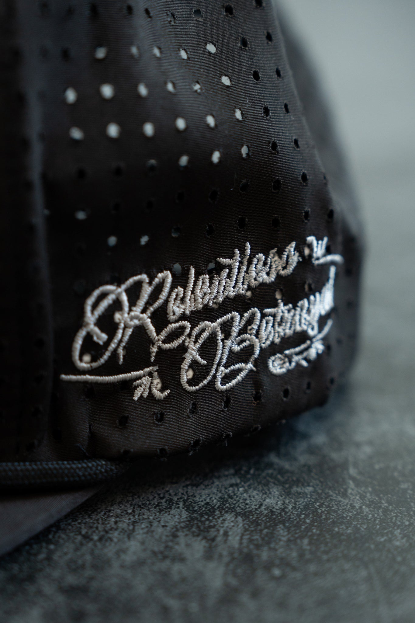 Close-up of the Black Flag Snapback by Relentless Betrayal, showcasing black perforated fabric embroidered with Relentless + Inspired in white script, highlighting the hats durability against a textured gray background.