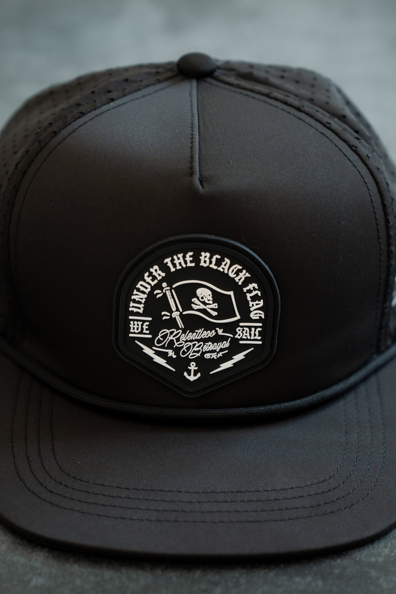 The Black Flag Snapback - Black by Relentless Betrayal features a circular Under the Black Flag logo with skull motifs, pirate-themed anchor details, and Relentless Beyond ETA phrase. It has a comfortable snapback closure and mesh back.