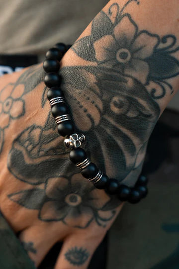 A hand adorned with floral and abstract tattoos showcases the Burn The Ships Bead Bracelet by Relentless Betrayal. The bracelet features black beads with a metal skull charm and spaced metal rings, all set against a soft, blurred background.