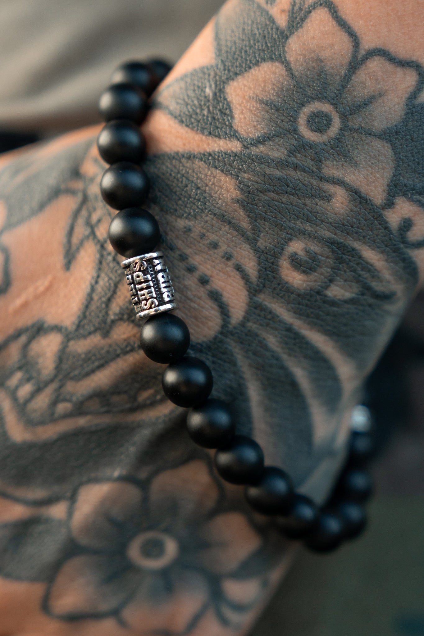 A close-up of a tattooed arm displays intricate floral designs alongside the Burn The Ships Bead Bracelet by Relentless Betrayal, featuring a silver Paris charm and a striking skull charm.