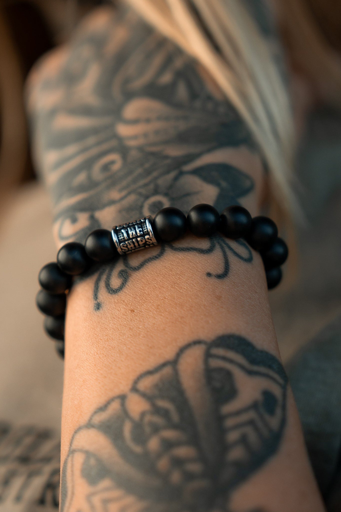 A tattooed forearm showcases Relentless Betrayals Burn The Ships Bead Bracelet with a black beaded band and metal accent, surrounded by intricate tattoo patterns.