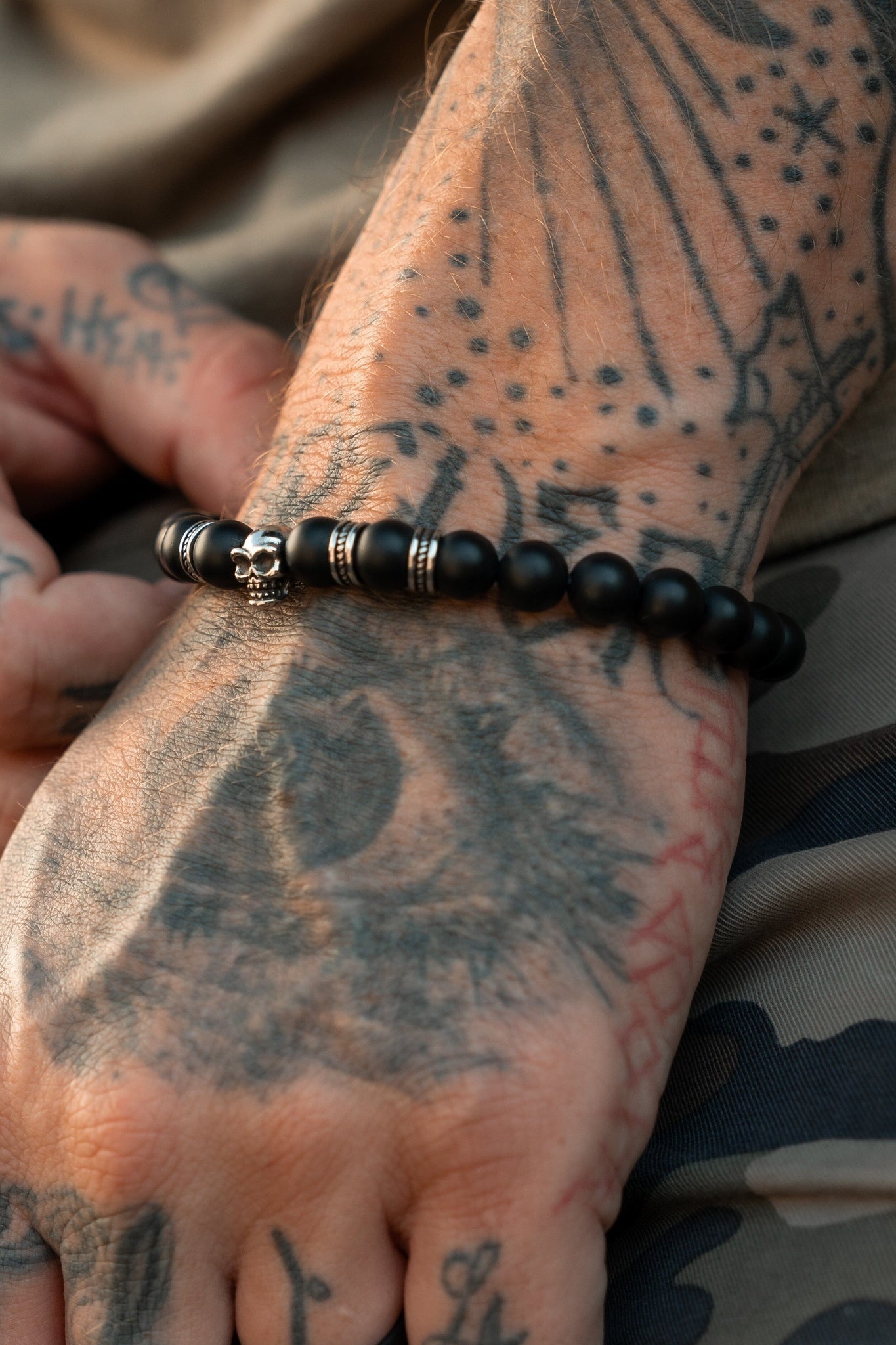 A tattooed hand, showcasing an eye design with abstract and geometric patterns, rests on a thigh. It wears the Relentless Betrayal Burn The Ships Bead Bracelet featuring a pirate skull. The person is dressed in camouflage pants.