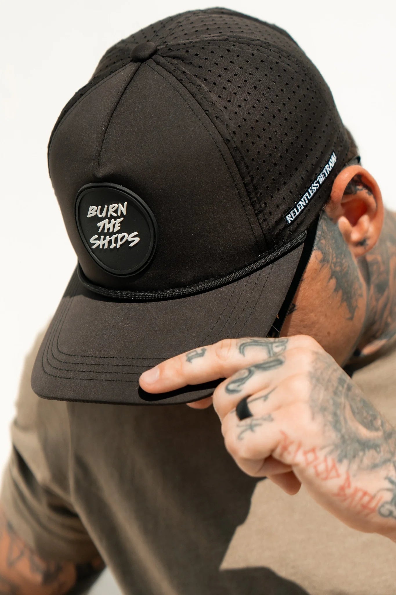 A person wearing a Burn The Ships Black PVC Snapback from Relentless Betrayal tilts their cap down, covering part of their face in a pirate-like manner, with tattoos on their hand and arm.