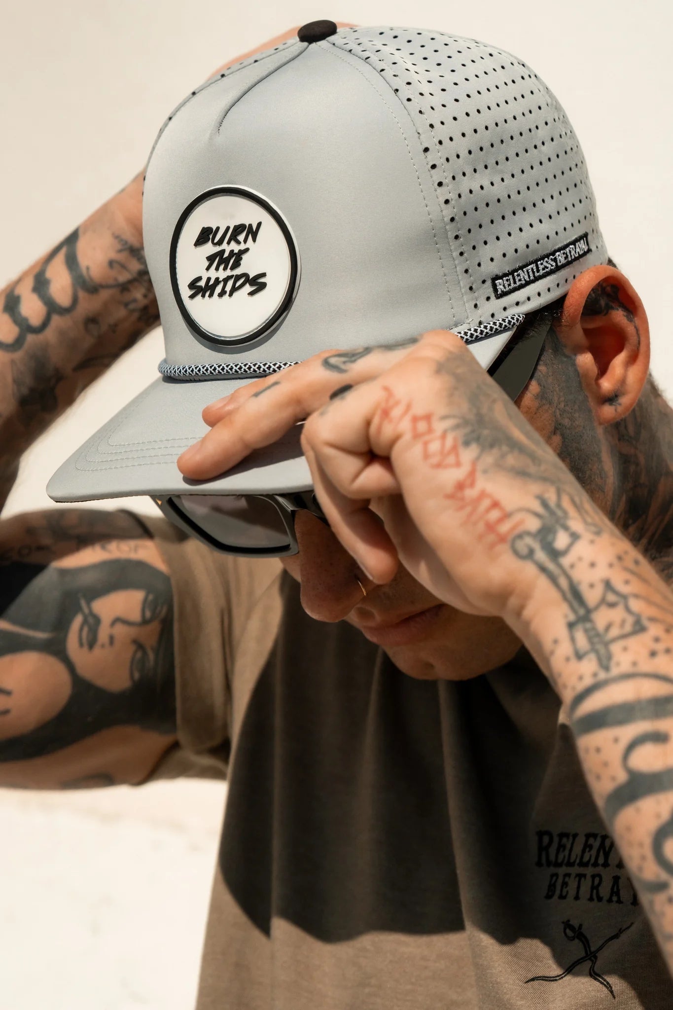 A tattooed person adjusts a Burn The Ships Cement PVC Snapback, paired with shades and a Relentless Betrayal shirt, embracing the pirate lifestyle against a simple light backdrop.