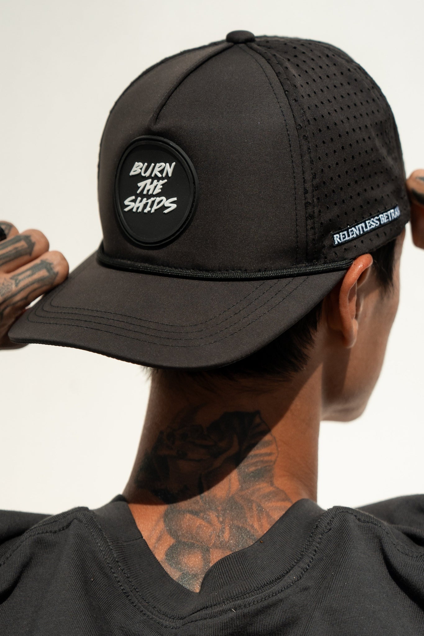 Wearing a Burn The Ships Black PVC Snapback by Relentless Betrayal, featuring BURN THE SHIPS on the front and RELENTLESS DEFENSE on the side, a person with tattooed neck and hands in a black shirt perfectly embodies a pirate lifestyle seen from behind.