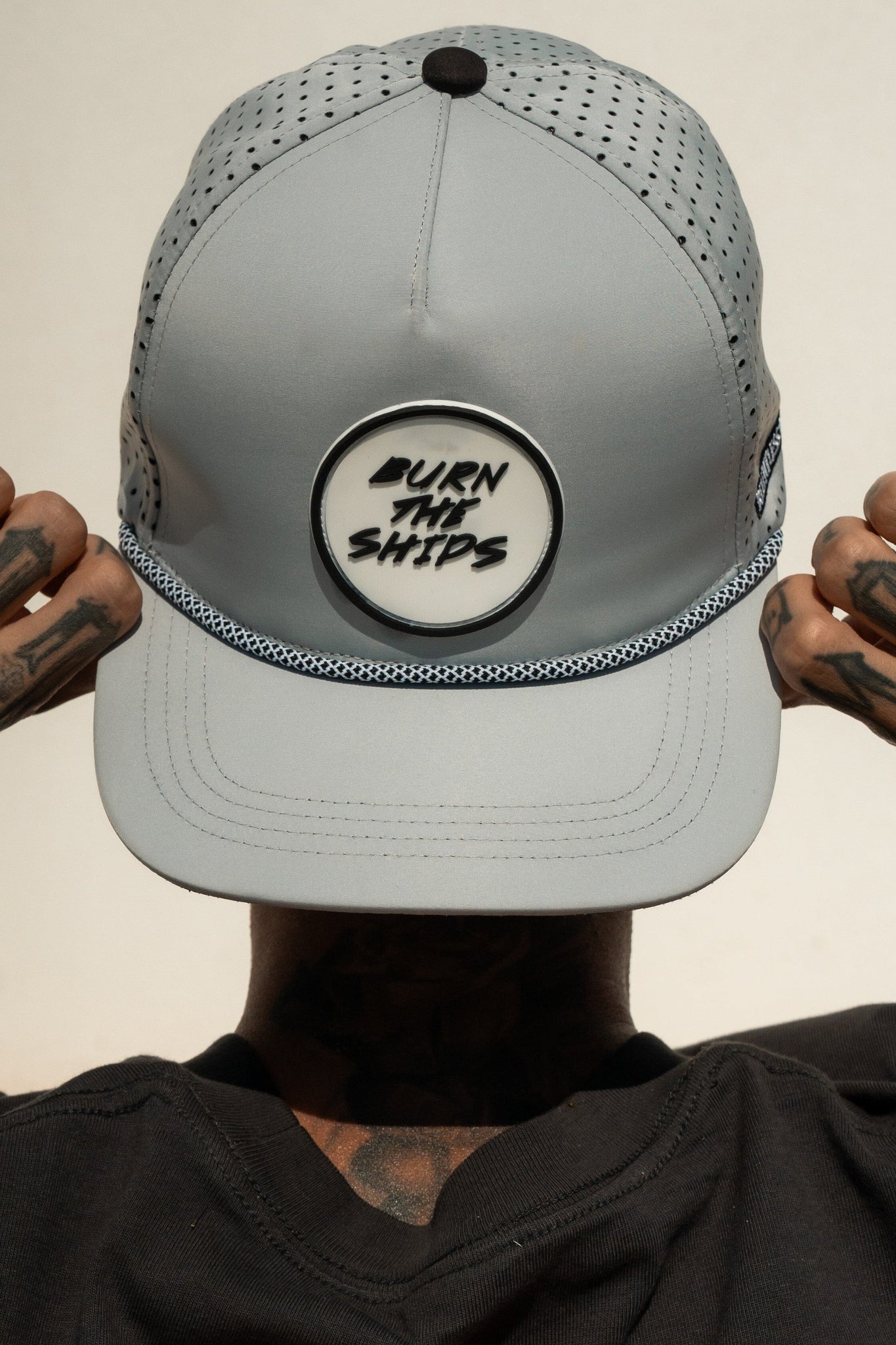 A person dons a Relentless Betrayal Burn The Ships Cement PVC Snapback, embodying the pirate spirit. The light gray cap boasts mesh sides, a textured base, and a water-resistant snapback for durability. Tattooed hands contrast with the plain backdrop.