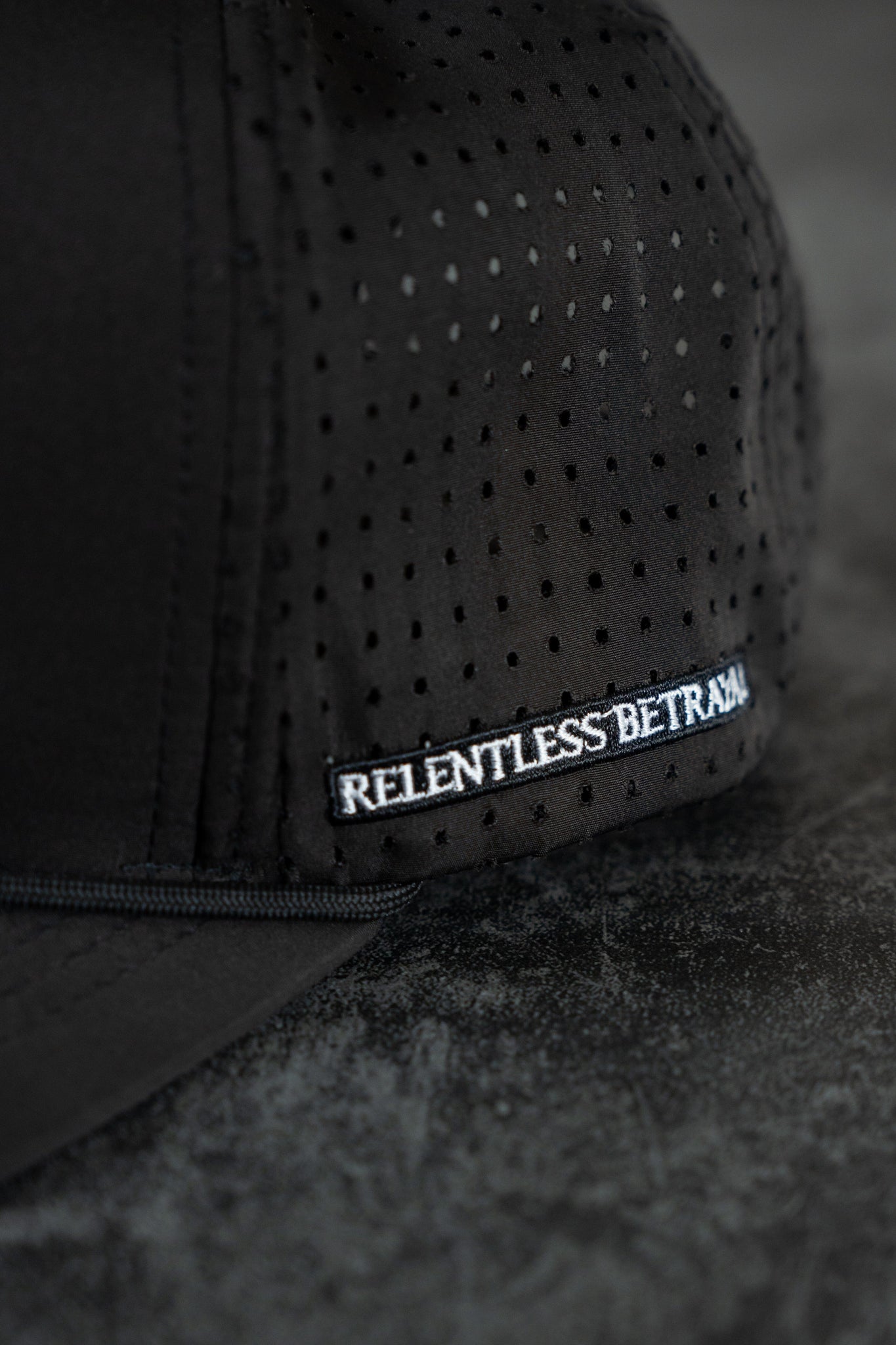 Close-up of the Burn The Ships Black PVC Snapback by Relentless Betrayal, featuring perforated fabric for ventilation and embodying the pirate lifestyle. A small side label reads RELENTLESS BETRAYAL in white letters against a dark, textured background.