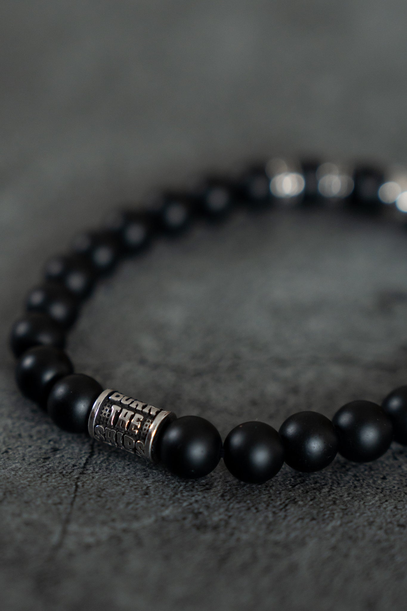 The Burn The Ships Bead Bracelet by Relentless Betrayal showcases black beads with a metallic, engraved bead and a skull charm, all against a textured dark gray backdrop.