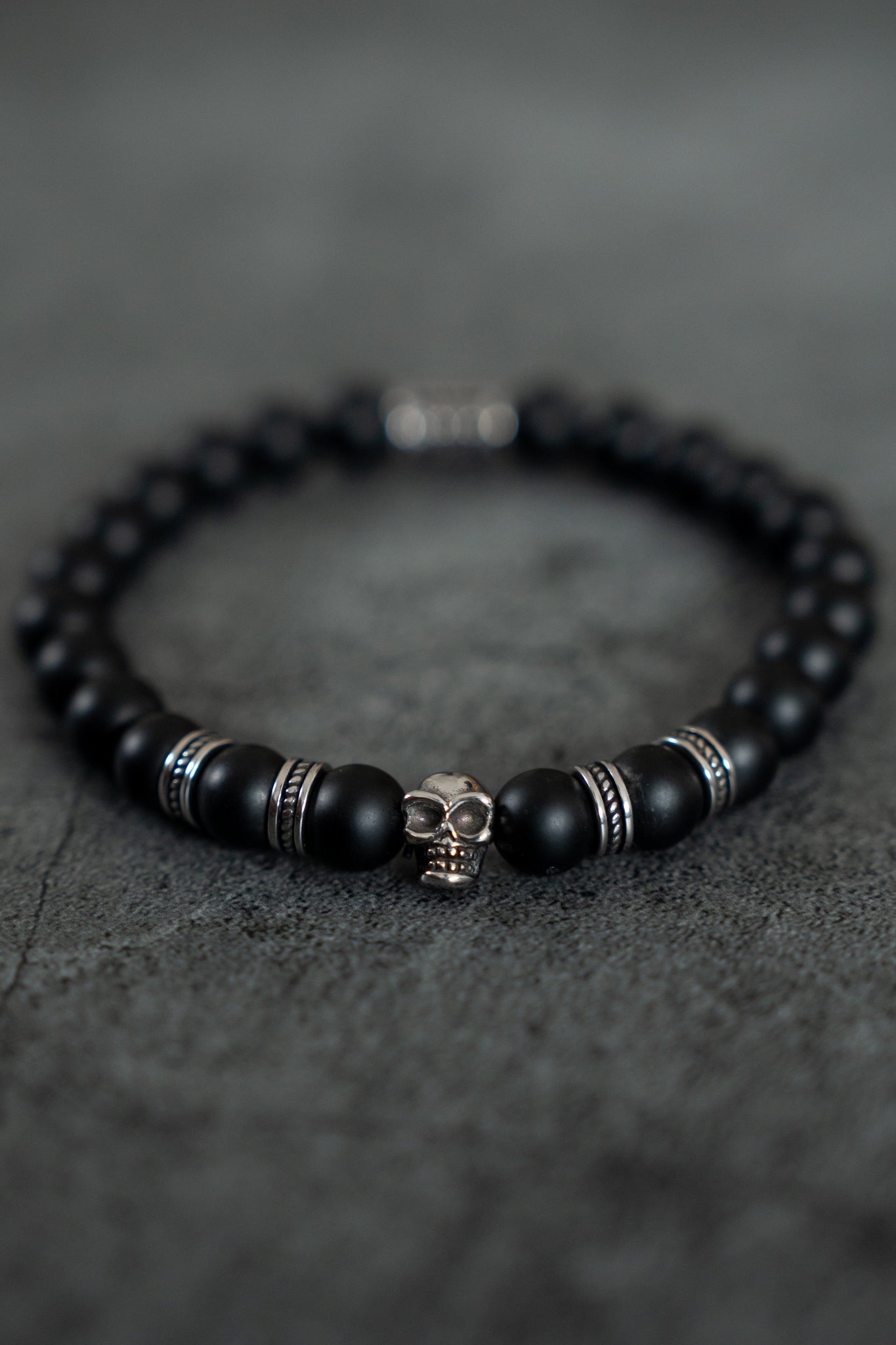 The Burn The Ships Bead Bracelet by Relentless Betrayal features a black beaded design with a striking silver skull charm, evoking pirate treasure. Accented with metallic highlights on dark gray, it embodies mystery and adventure.
