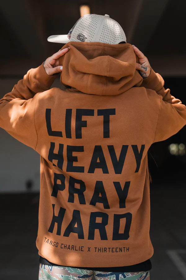 A person wears a tan Lift Heavy. Pray Hard. - Hoodie by Tango Charlie Apparel, hood up and hands raised to show the bold black "LIFT HEAVY PRAY HARD" print on the back, paired with a light-colored cap.