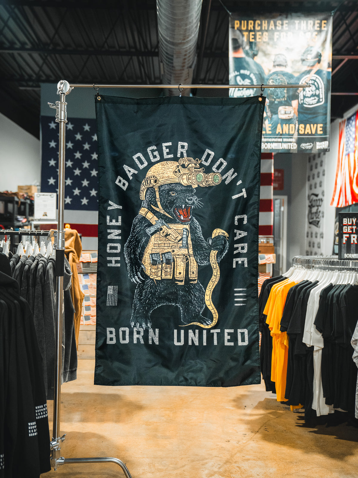 A Honey Badgerin' Flag from Born United Private Label hangs in a clothing store, showing an armored badger with gold gear and the text "HONEY BADGER DON’T CARE" and "BORN UNITED." T-shirts and American flags are seen behind it.
