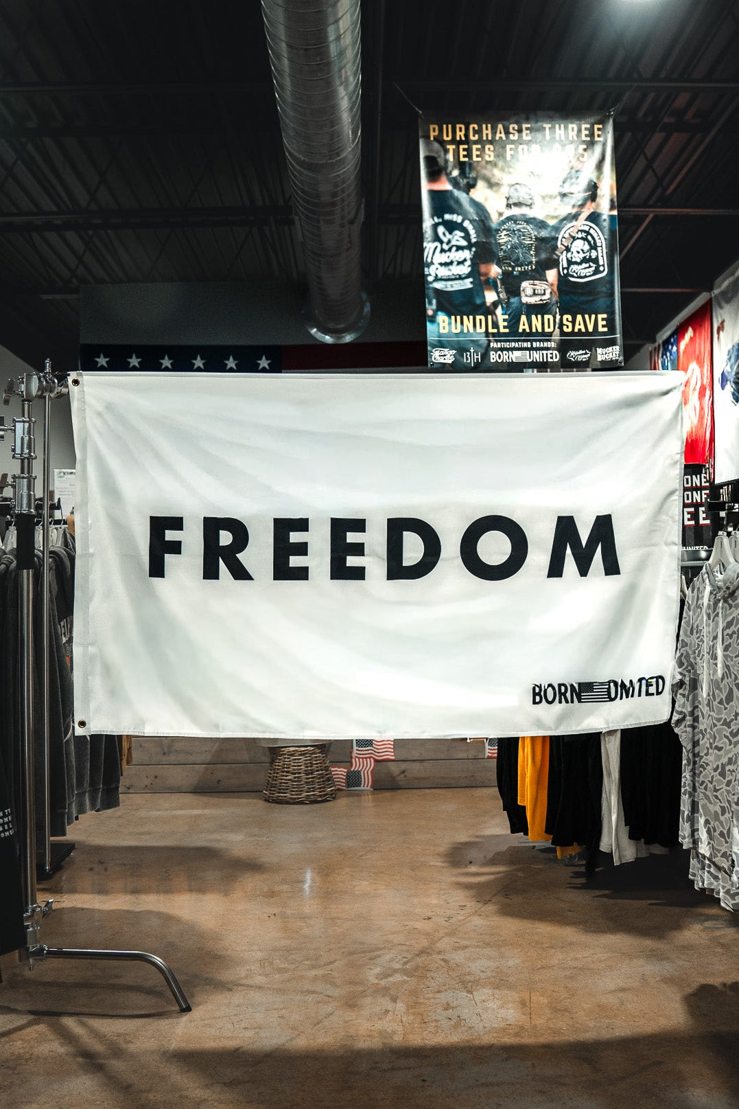 A large white Freedom Flag by Born United Private Label with bold black "FREEDOM" lettering hangs between clothing racks in a store, reflecting patriotic values. Various shirts and posters can be seen in the background.