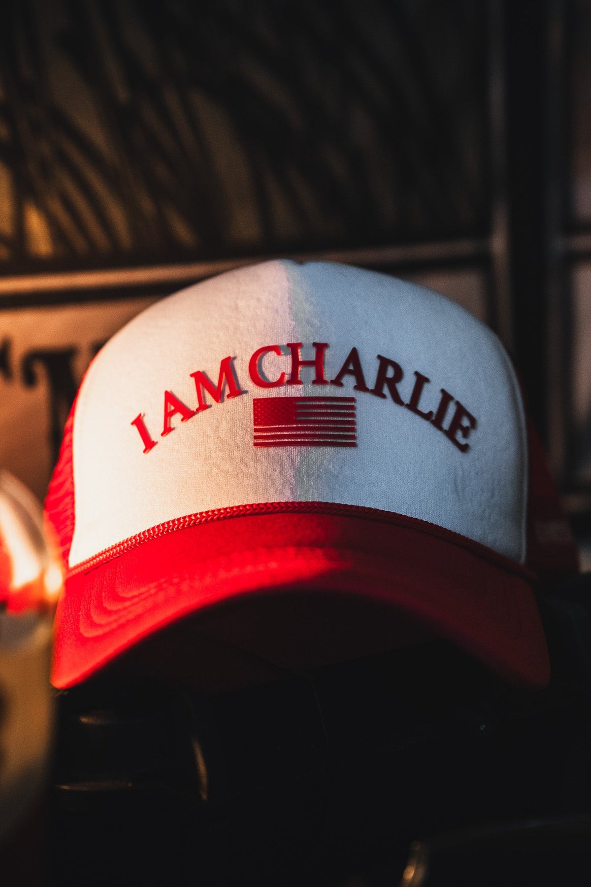 The Born United Private Label I Am Charlie Red Trucker Snapback features a red and white 5-panel curved brim, "I AM CHARLIE" text, and a stylized American flag on the front, shown in focus resting indoors against a dark background.